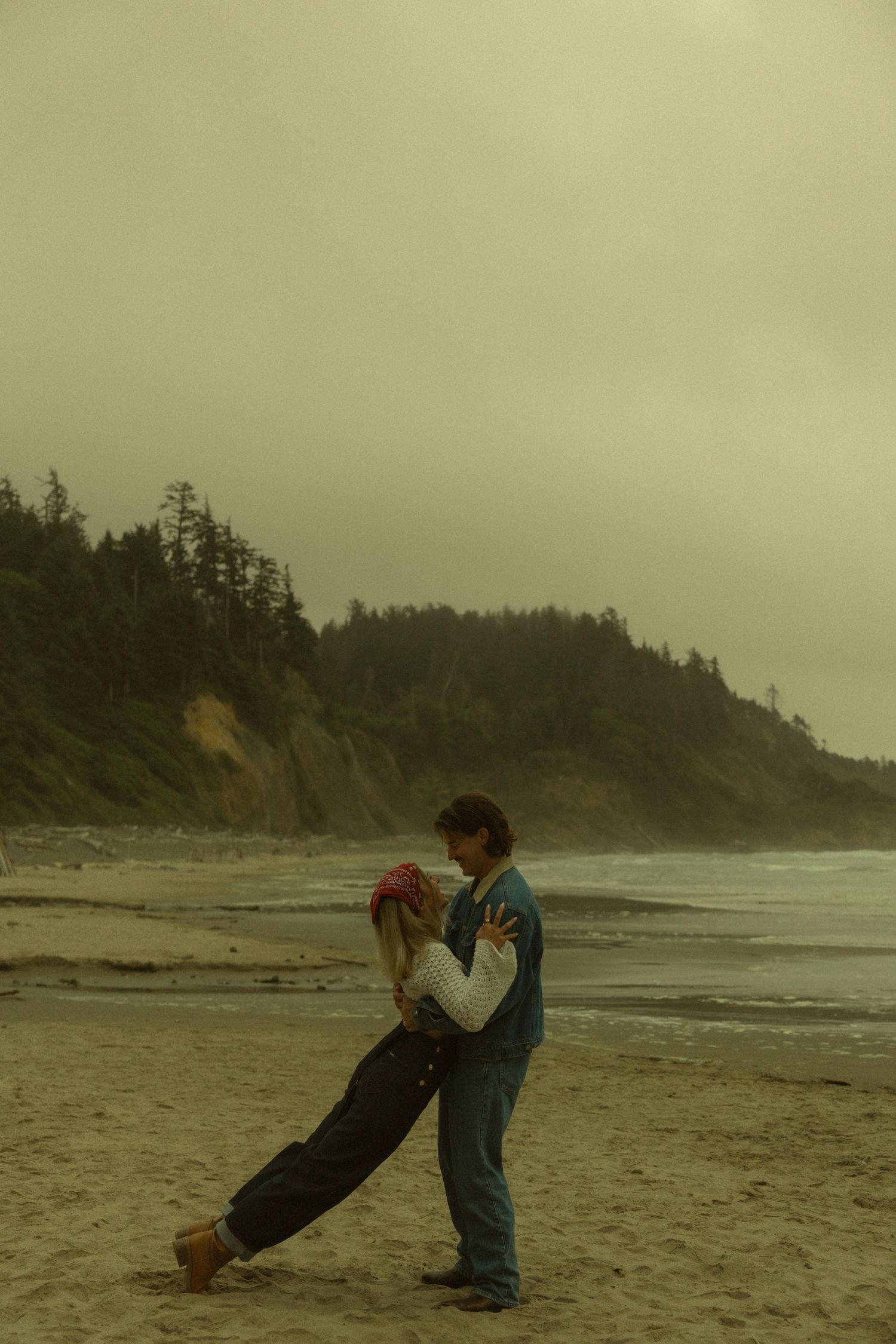 A couple dances romantically on a misty beach with a tree-lined coastal bluff in the background.