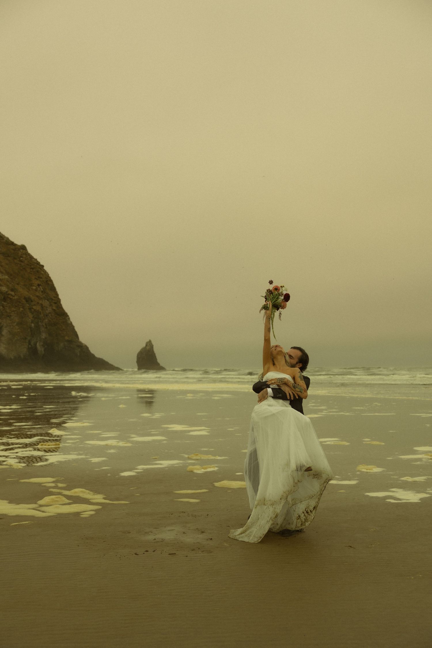 Bride and groom celebrating on misty Cannon Beach, lifting bouquet with Haystack Rock in background.