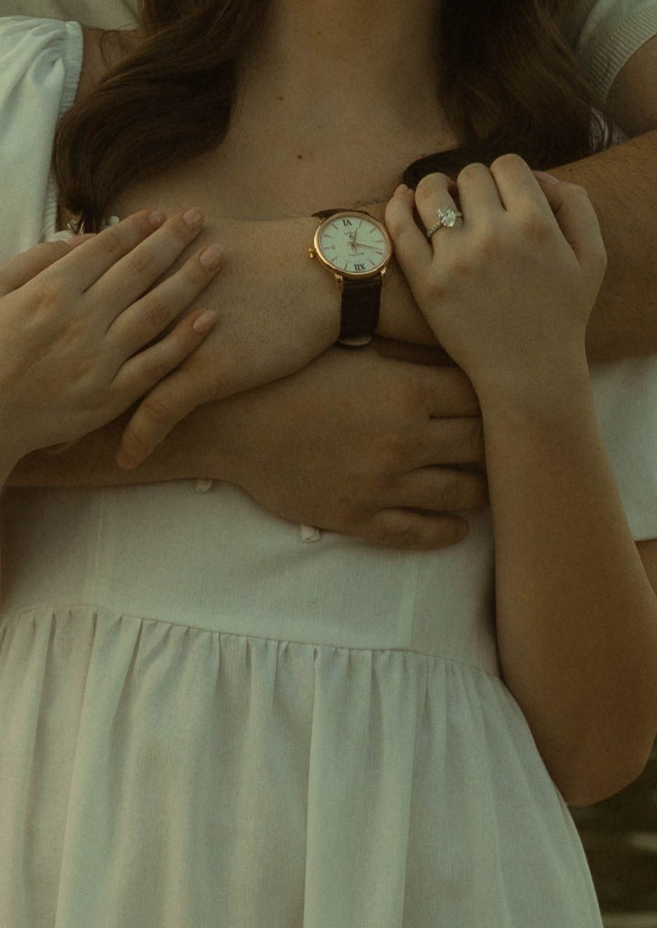 Couple embracing, woman in white dress wearing a gold watch and diamond engagement ring.