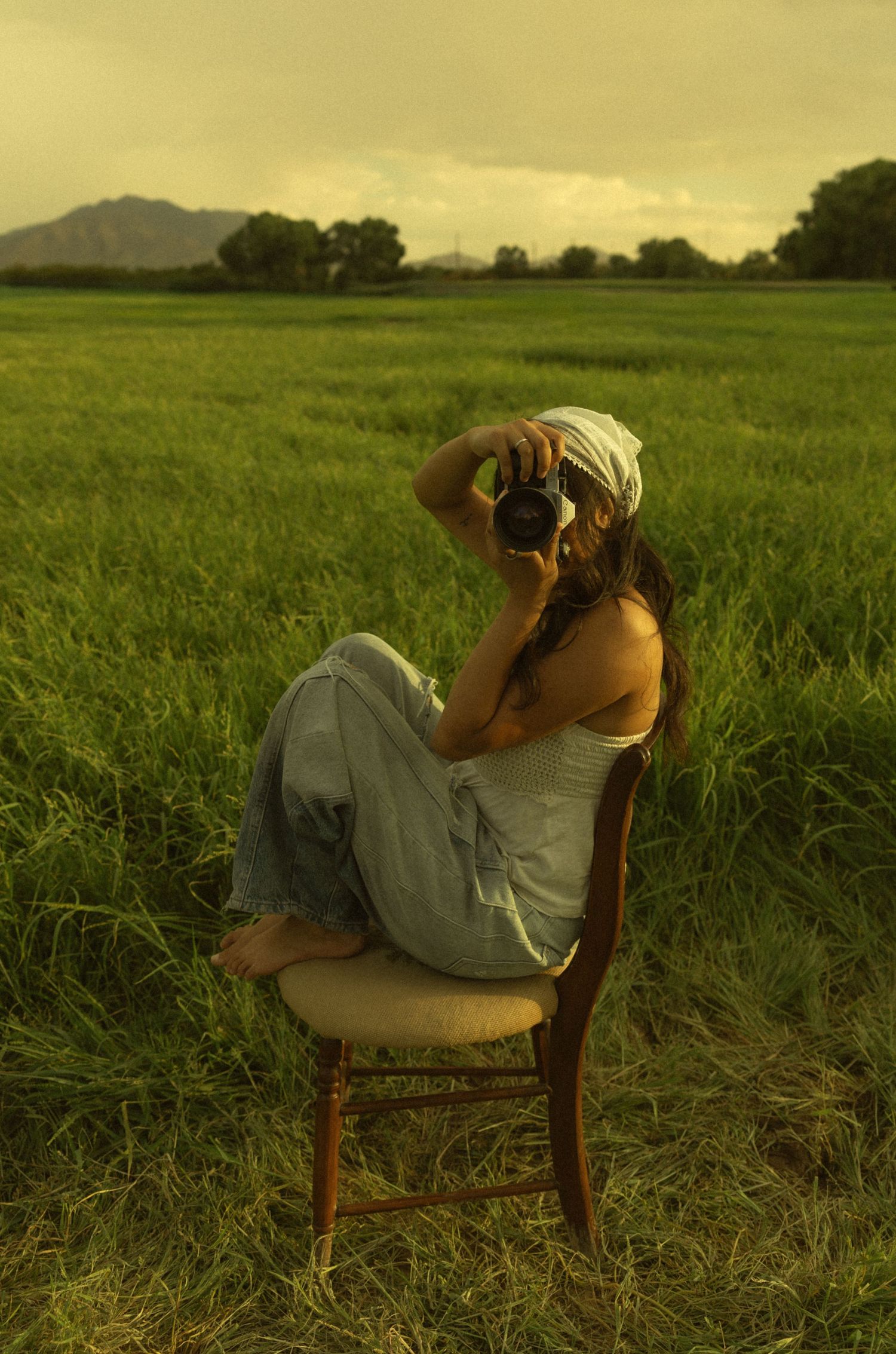 Woman sitting on a wooden chair in a green field, holding a camera up to her face on a cloudy day.