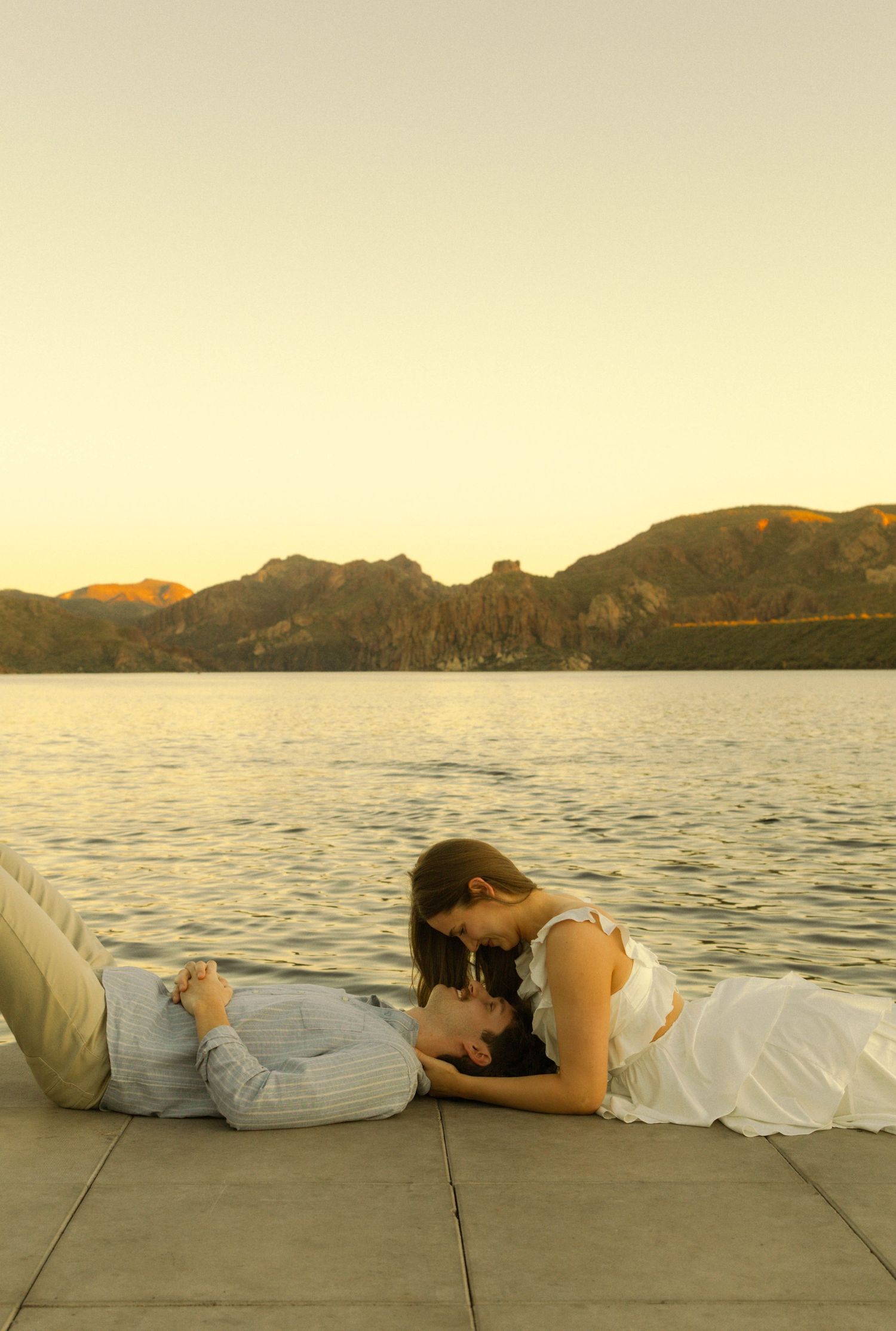 A couple relaxes on a waterfront dock at sunset with mountains visible across the lake.