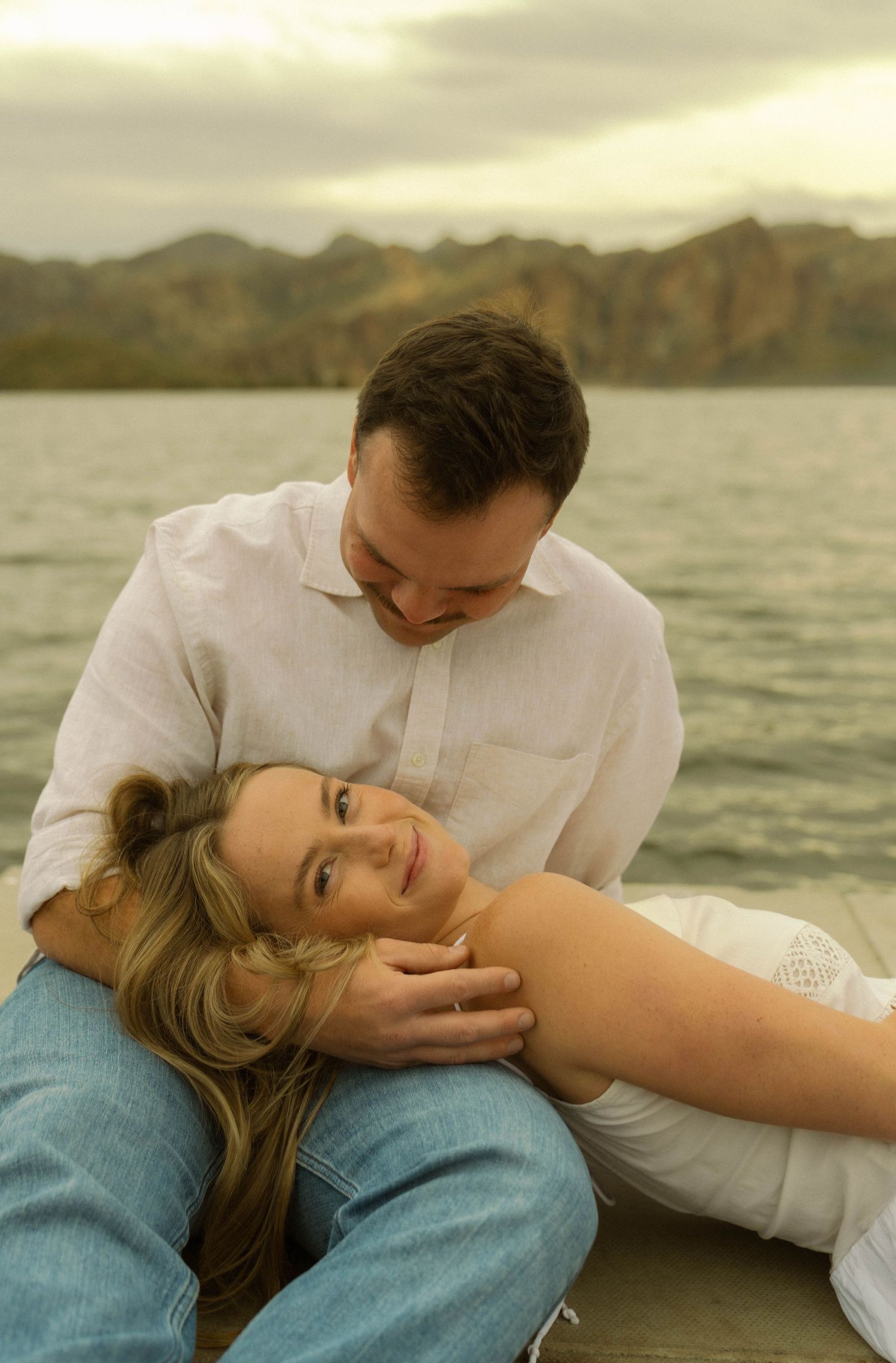 A couple shares an intimate moment while lying down near a scenic lake with mountains.