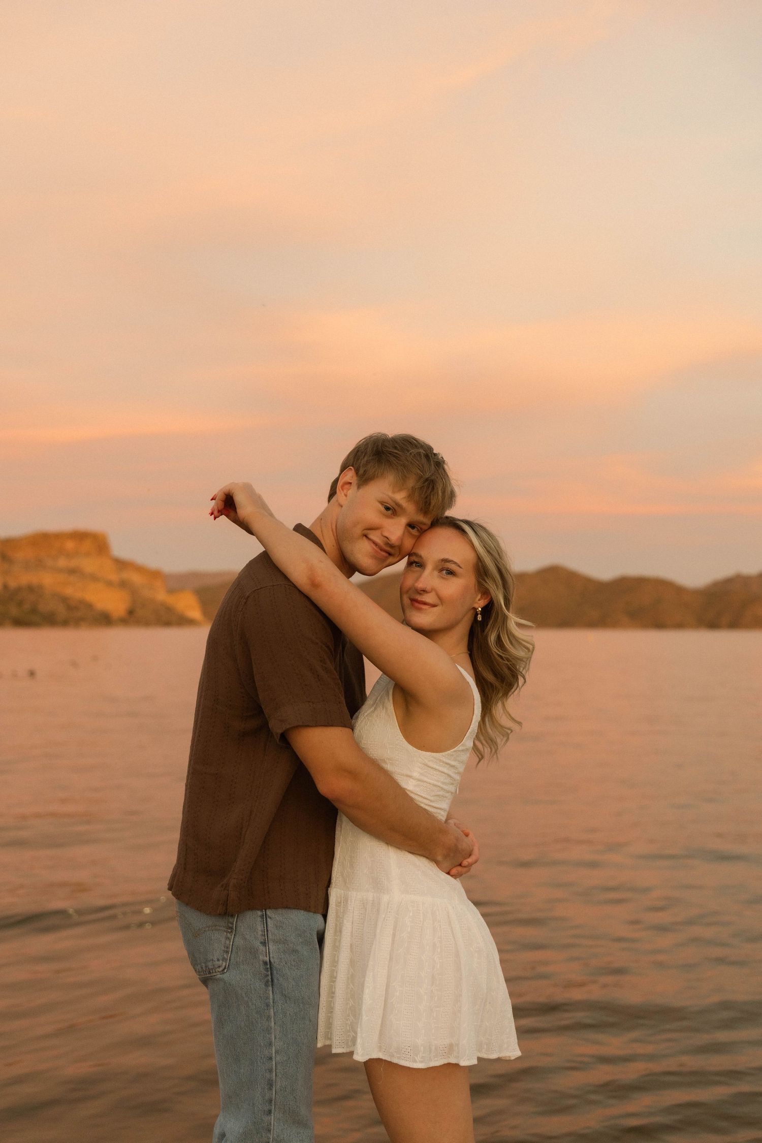 A couple dances playfully on a dock during a golden sunset overlooking a mountain lake.