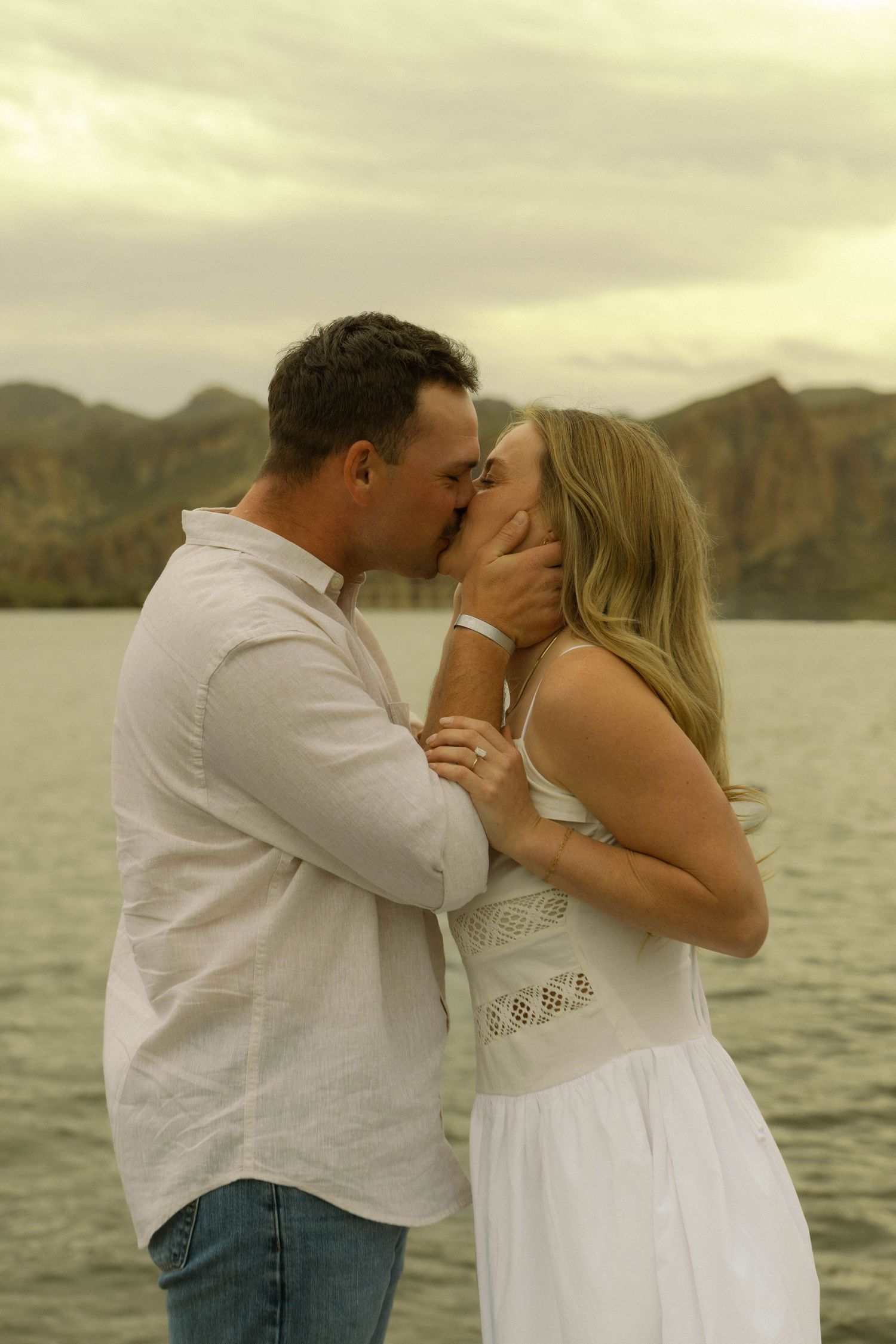 A romantic couple shares a tender kiss by the water with mountains in the background at sunset.