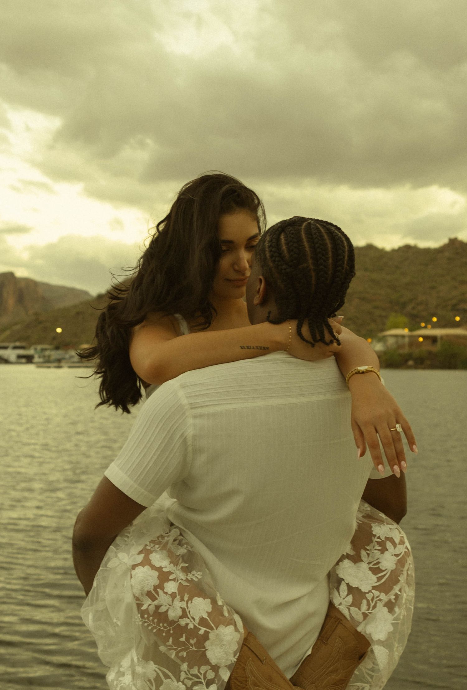 Couple embracing romantically by a lake at dusk, woman in white floral dress being lifted by man with braided hair.