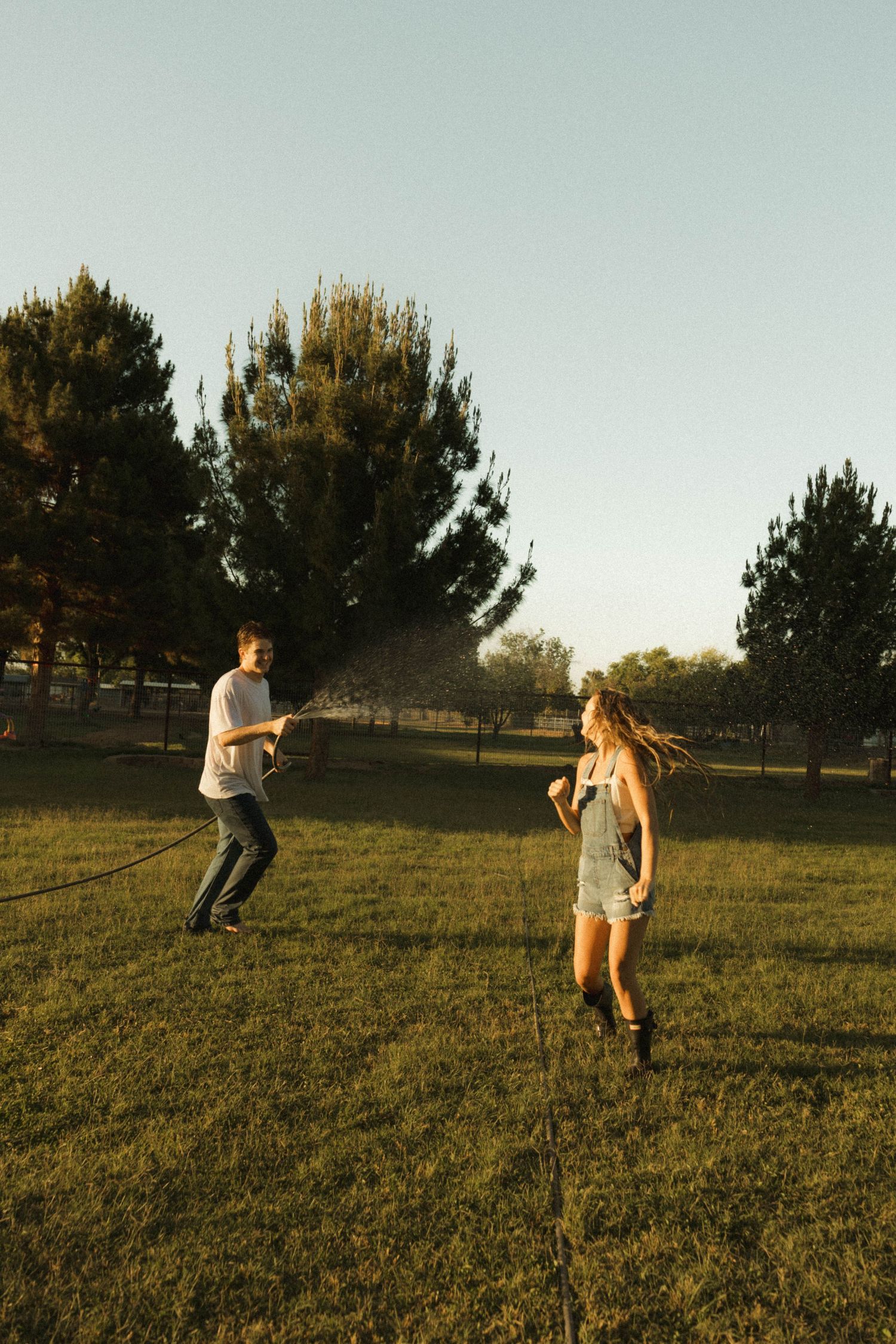 Man spraying woman with a garden hose as she laughs and runs across a sunny green field.