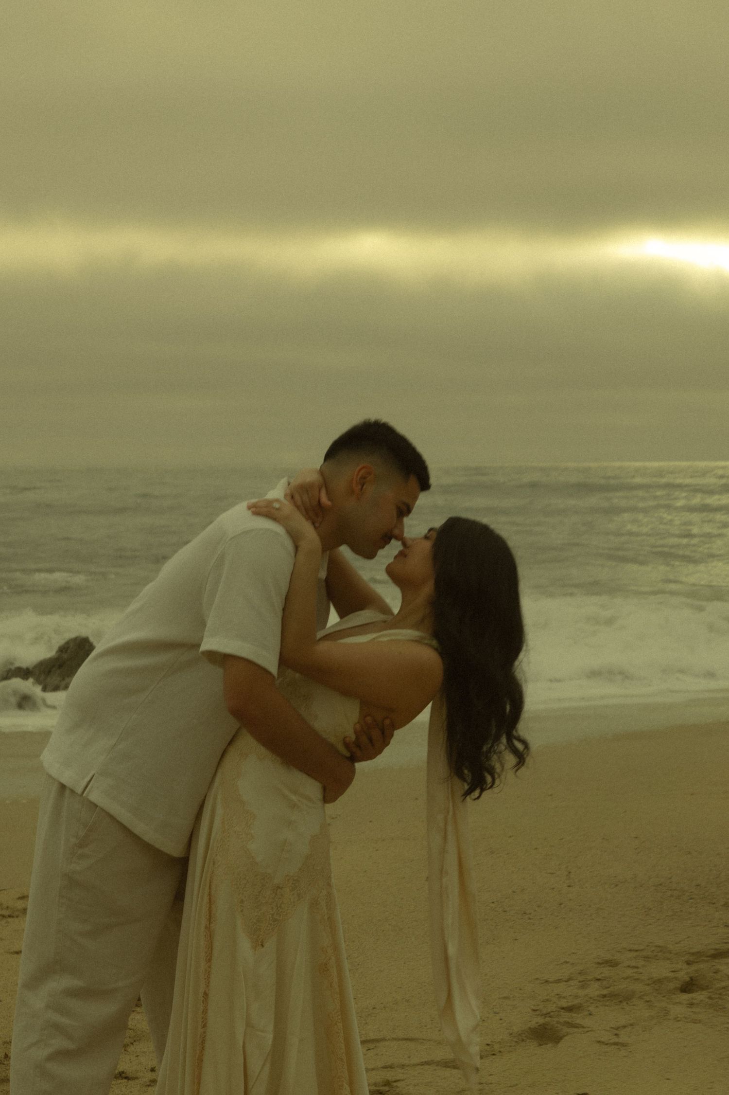 Couple sharing a romantic moment on a beach at dusk with golden cloudy skies and ocean waves.