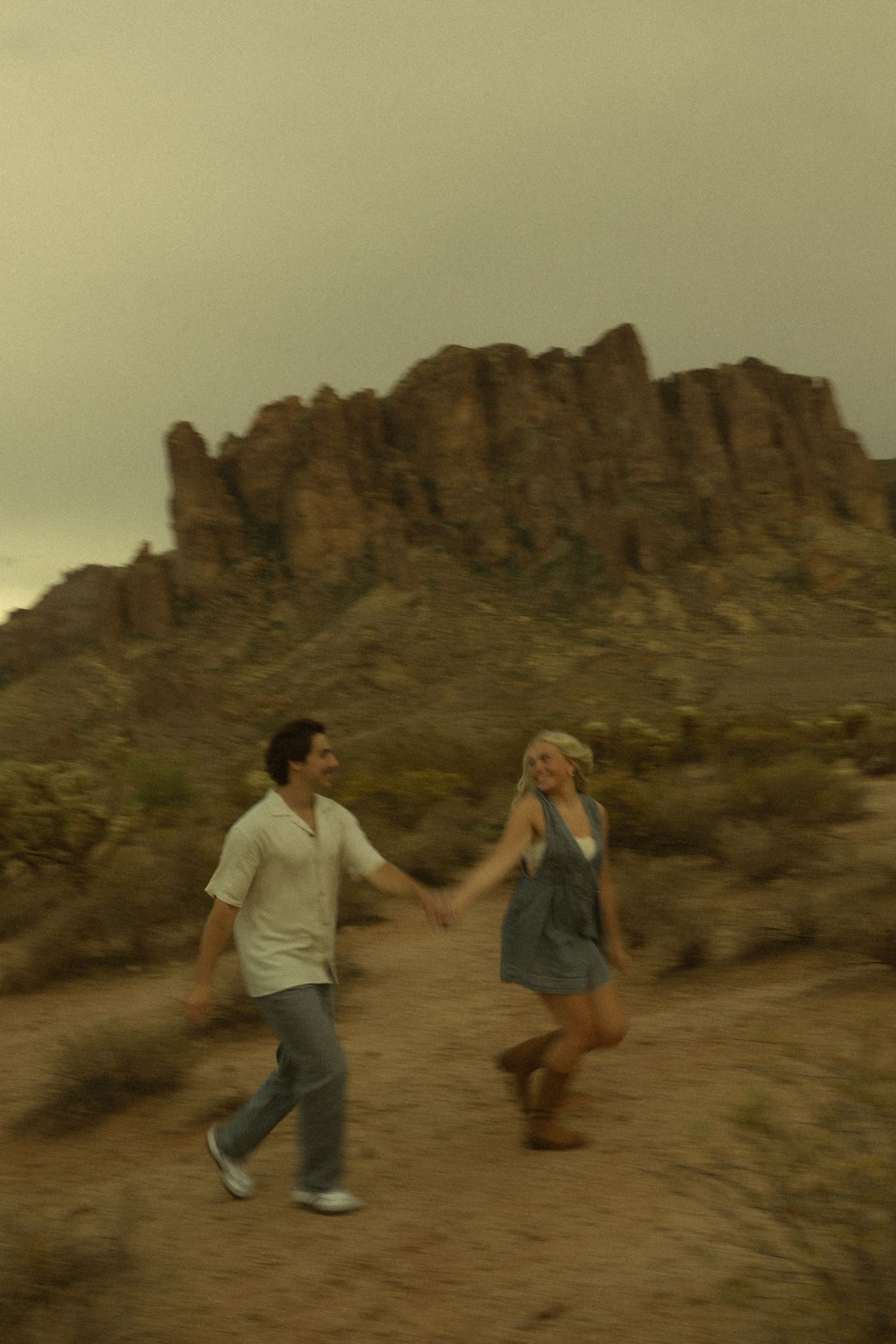 A couple holds hands and walks through a desert landscape with rugged rocky mountains in the background.