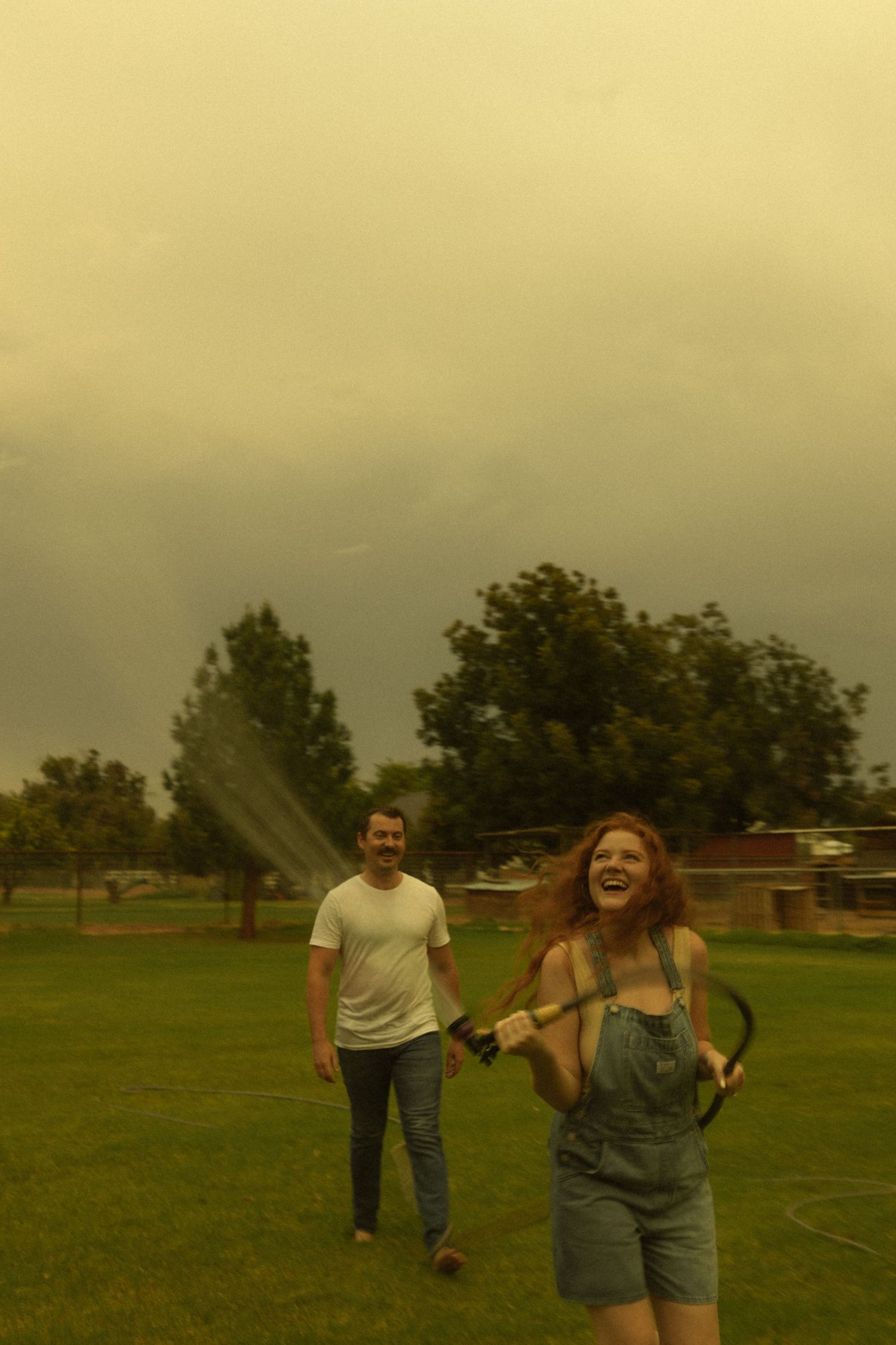 A laughing redhead woman in overalls holds a garden hose while a man in a white t-shirt smiles behind her outdoors.