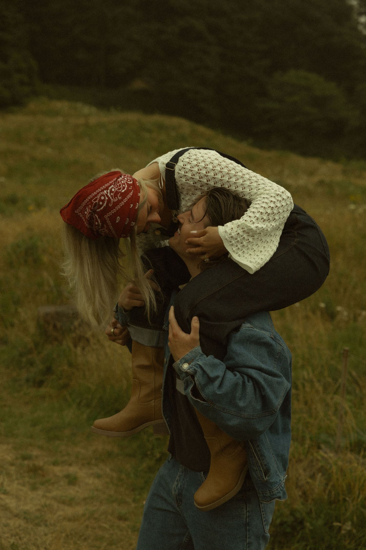 Couple embracing outdoors, woman in red bandana and boots being lifted by partner in denim jacket in grassy field.