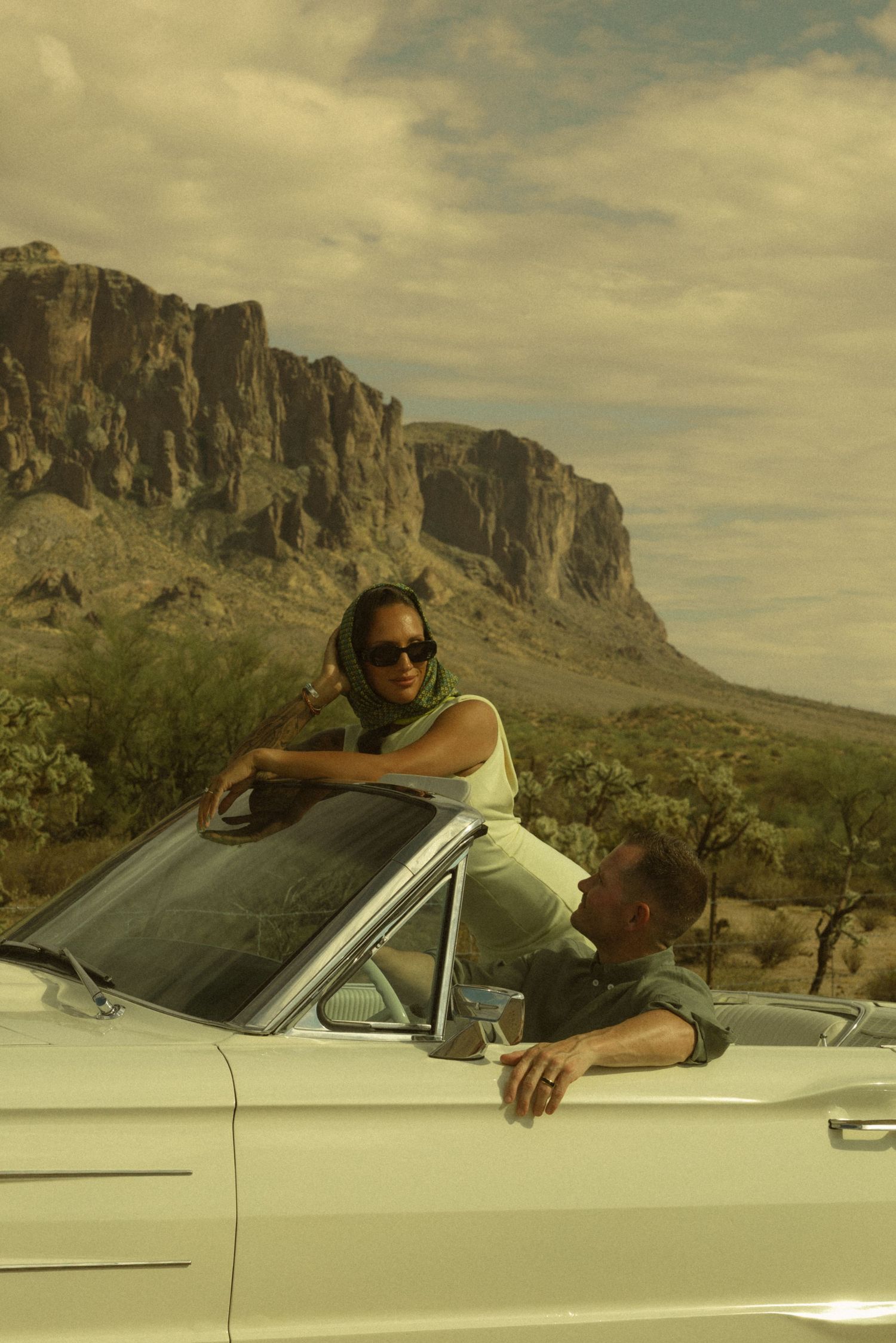 Couple in a white convertible car with dramatic desert mountains and cloudy sky backdrop.