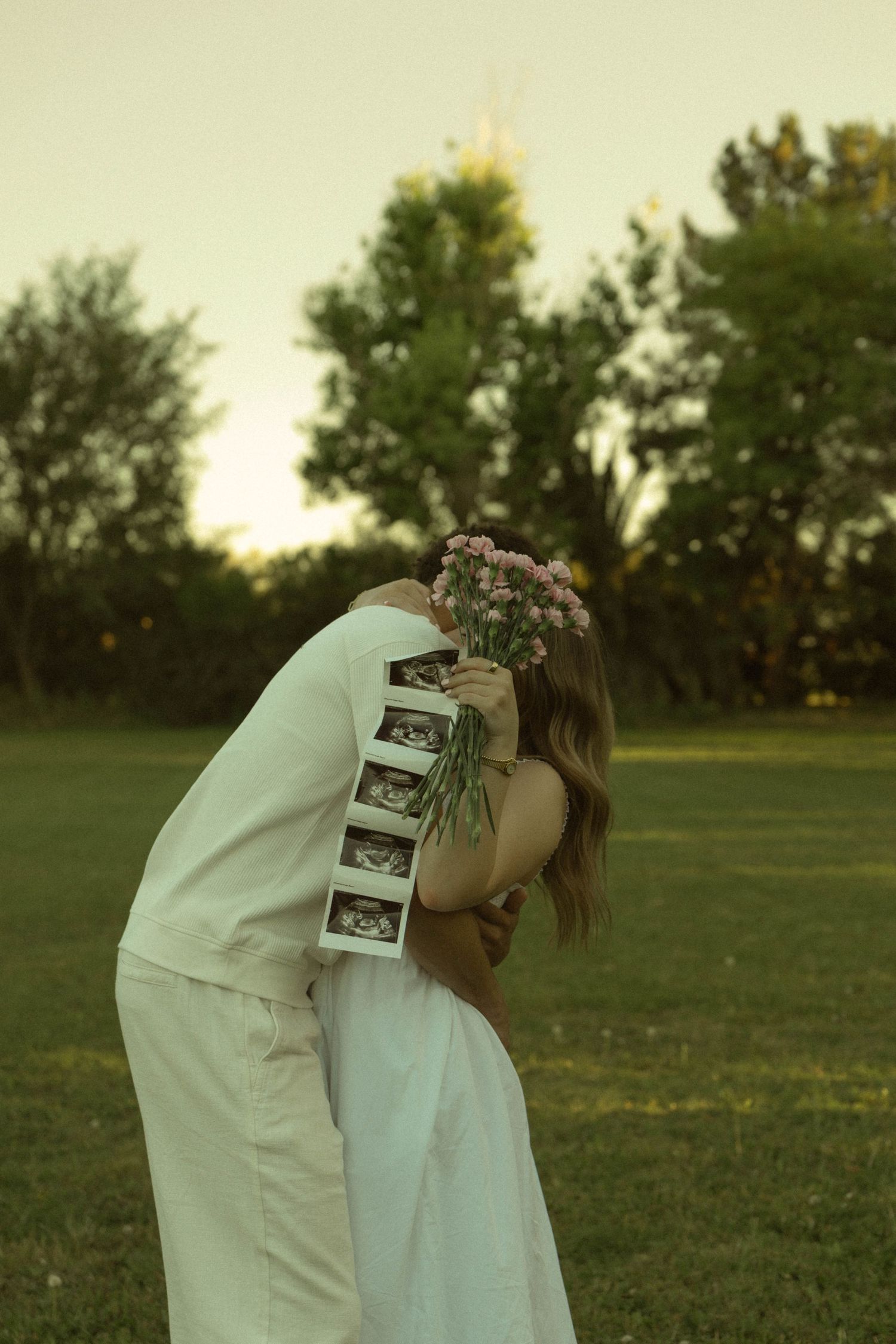 Couple kissing behind ultrasound photos and pink flowers during outdoor pregnancy announcement shoot.
