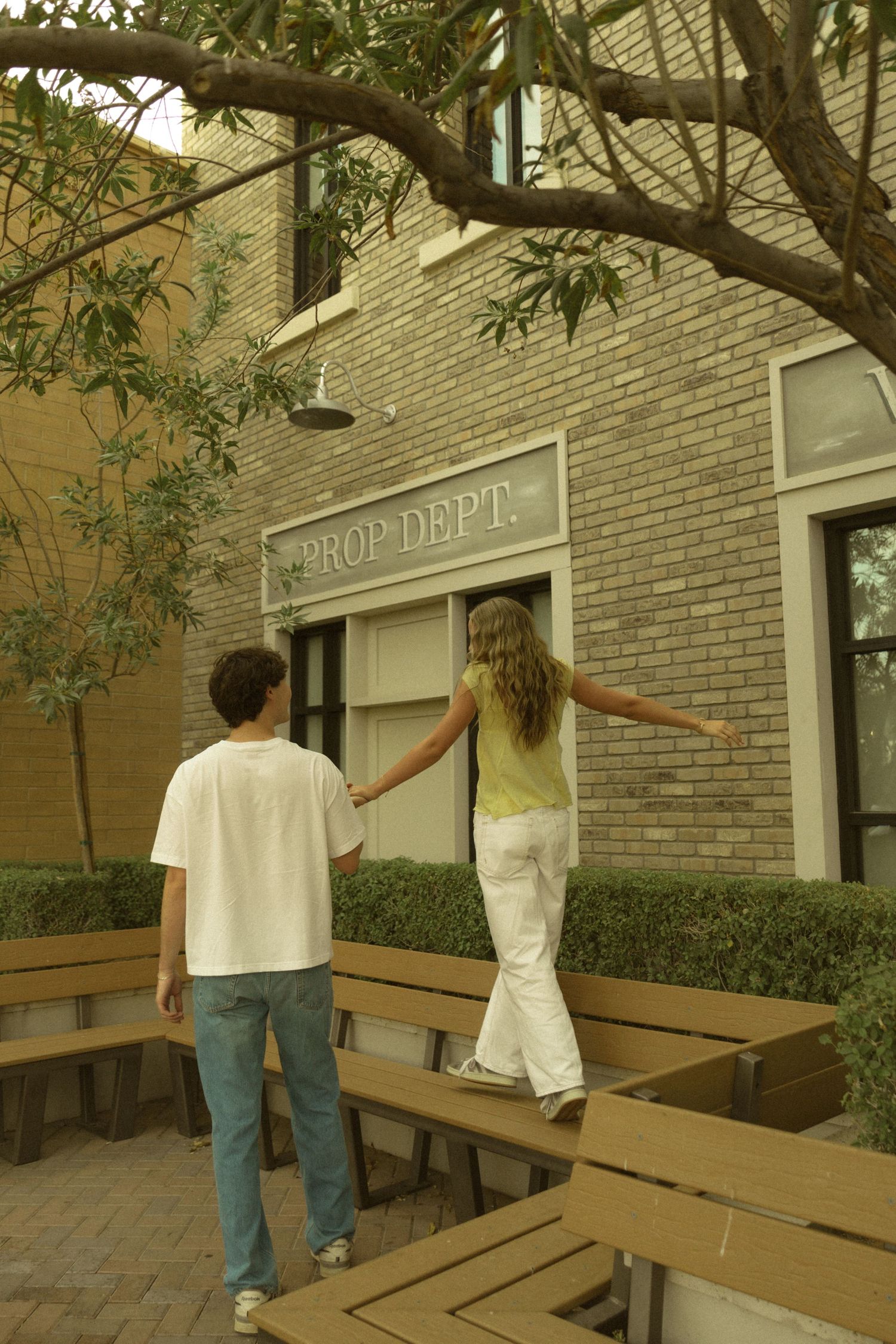 Young couple holding hands outside Prop Dept building, girl balancing on bench while boy steadies her.