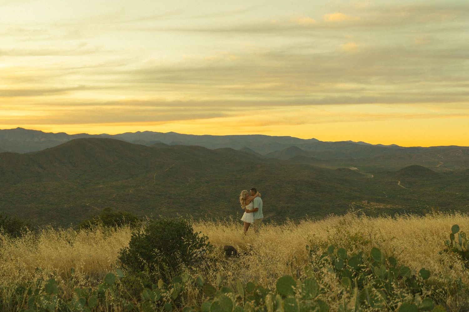 Couple embracing on a golden hillside at sunset with rolling mountains and dramatic sky in the background.