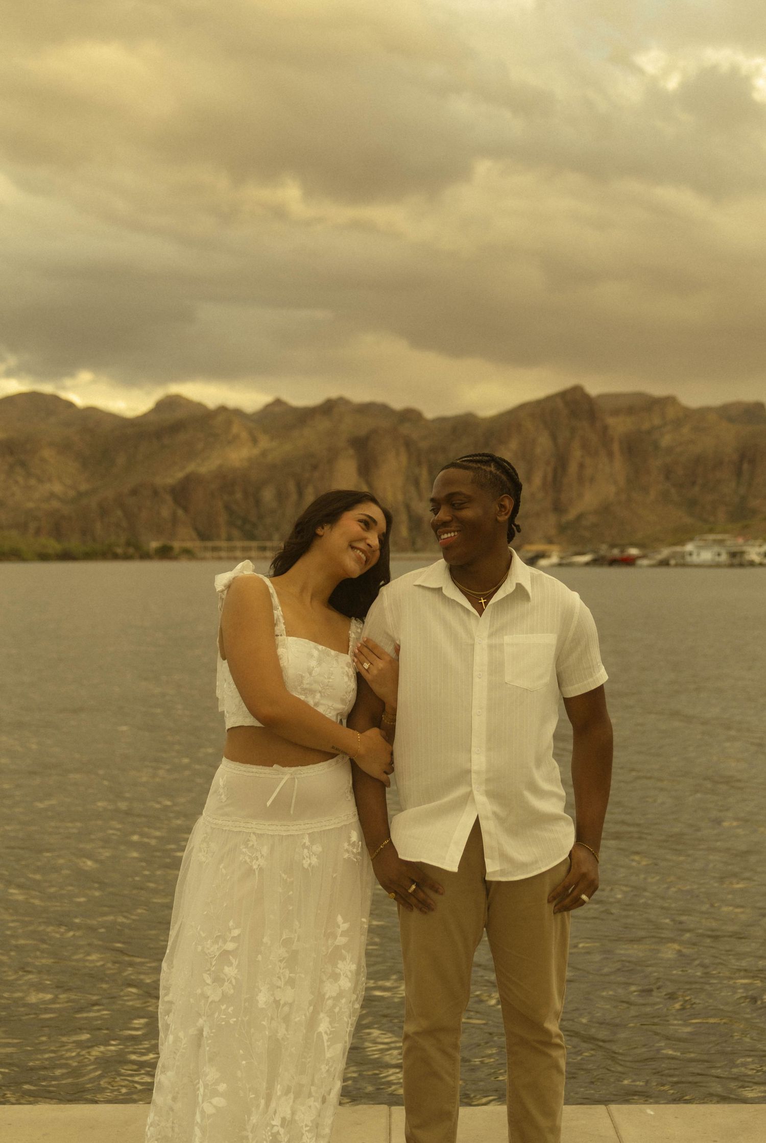 Couple in white outfits smiling by a lake with mountain backdrop at golden hour.