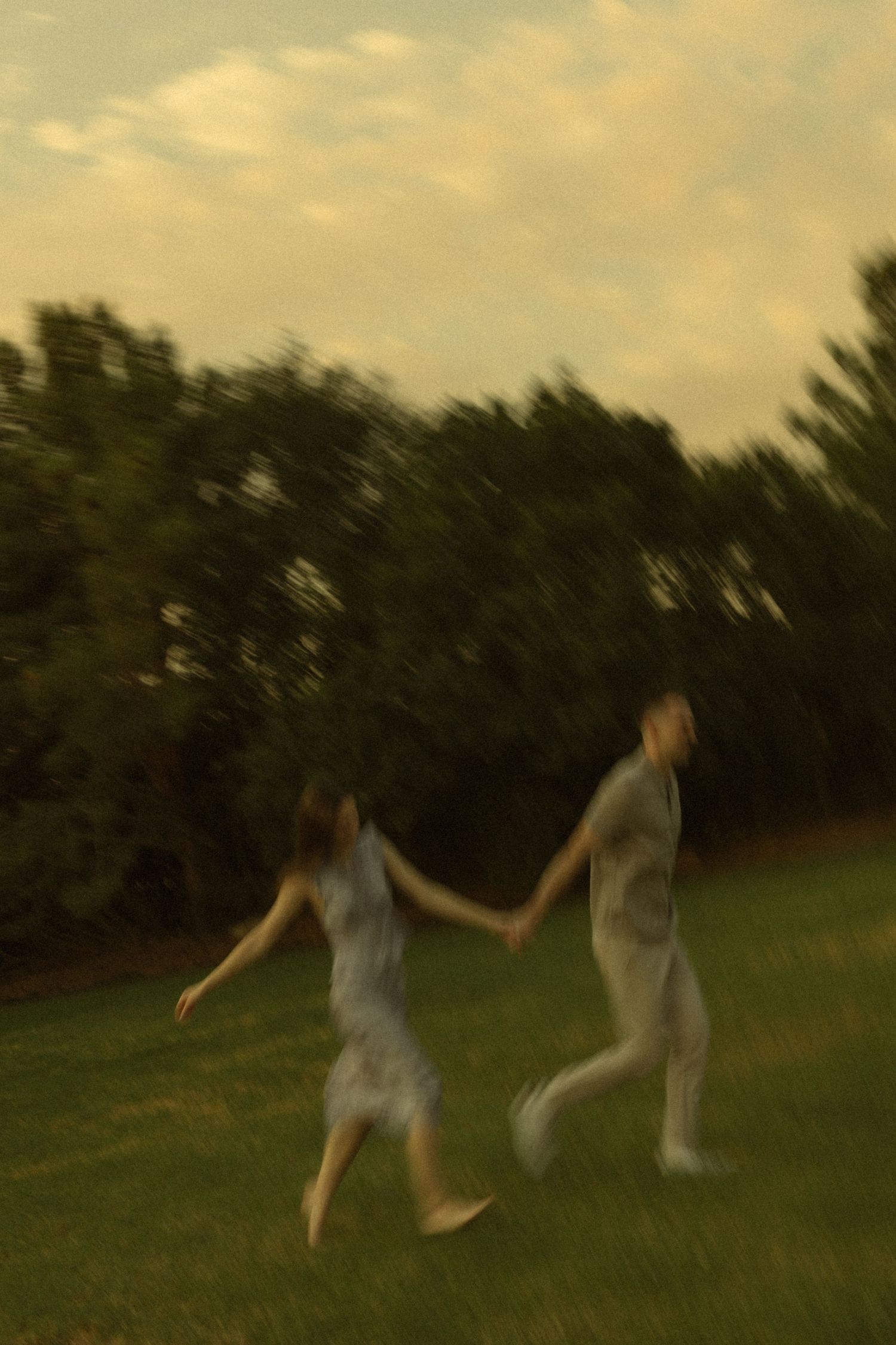 Blurry motion photo of a couple holding hands and running together on green grass with trees in the background.