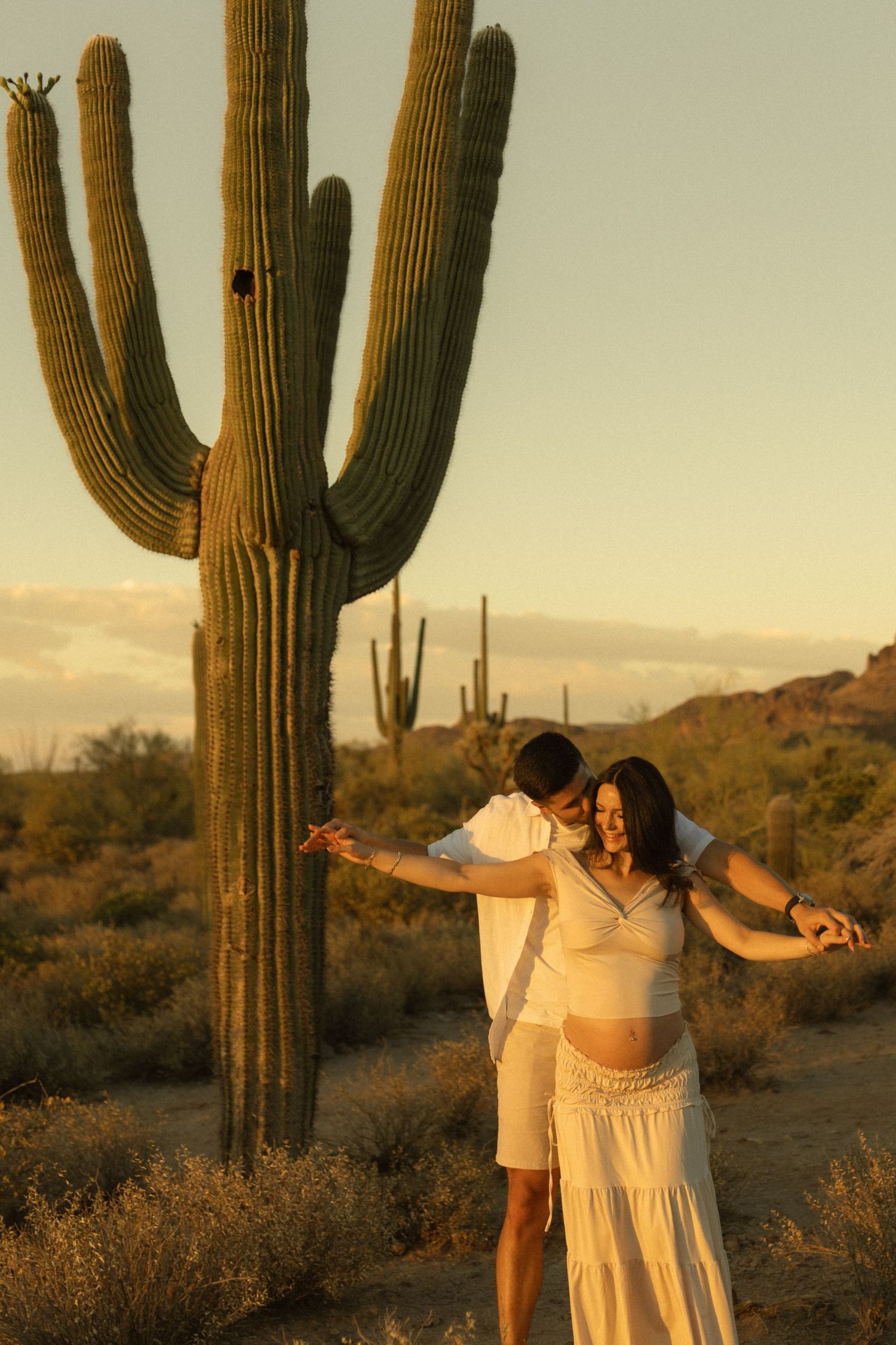 Pregnant couple dancing near saguaro cactus in Arizona desert at golden hour sunset.