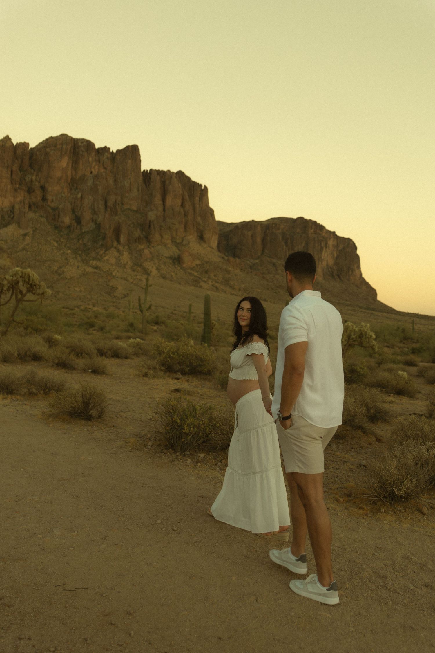 Pregnant couple holding hands on desert trail with Superstition Mountains at sunset.