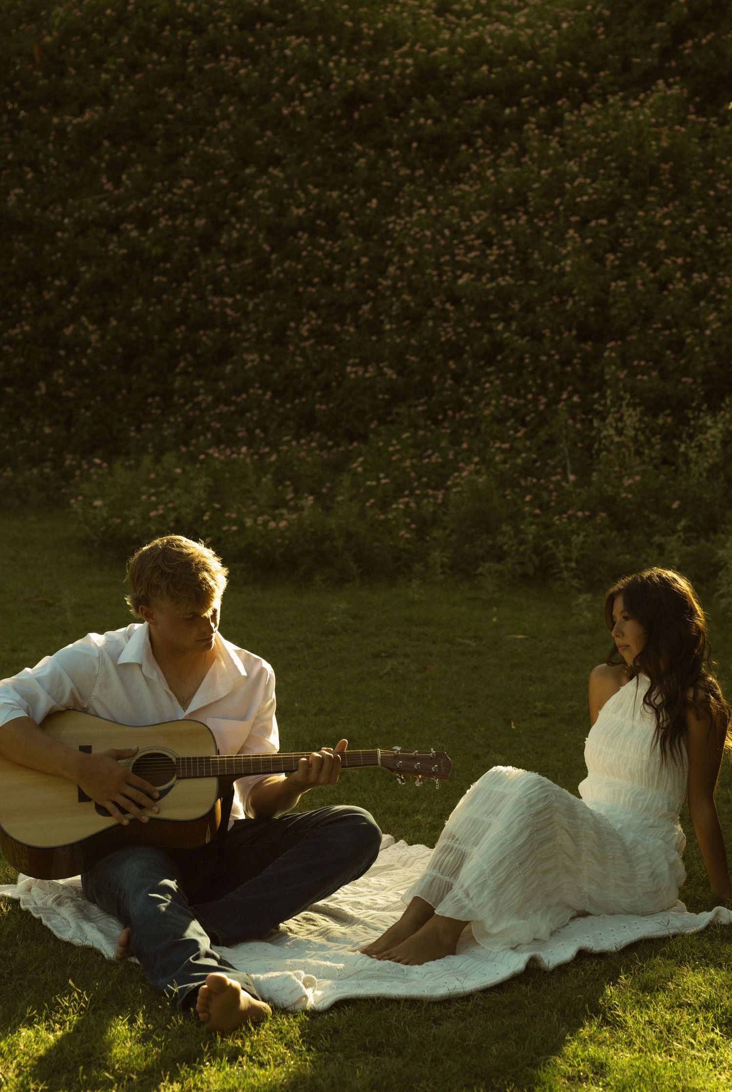 Couple sharing a romantic picnic on grass, man playing acoustic guitar for woman in white dress at sunset.