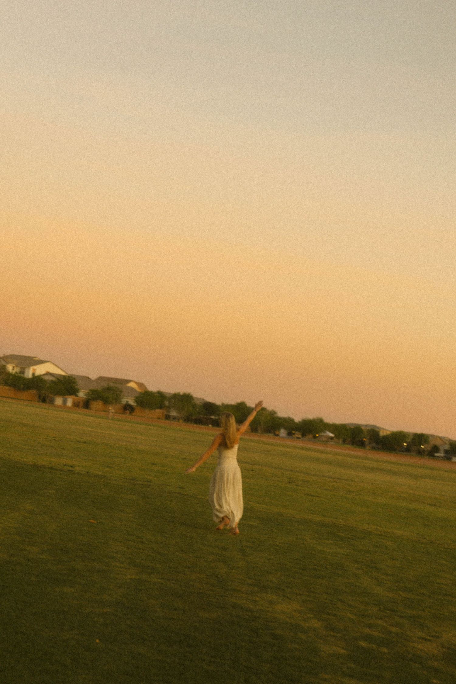 Woman in white dress standing in open field with arms raised during golden sunset.
