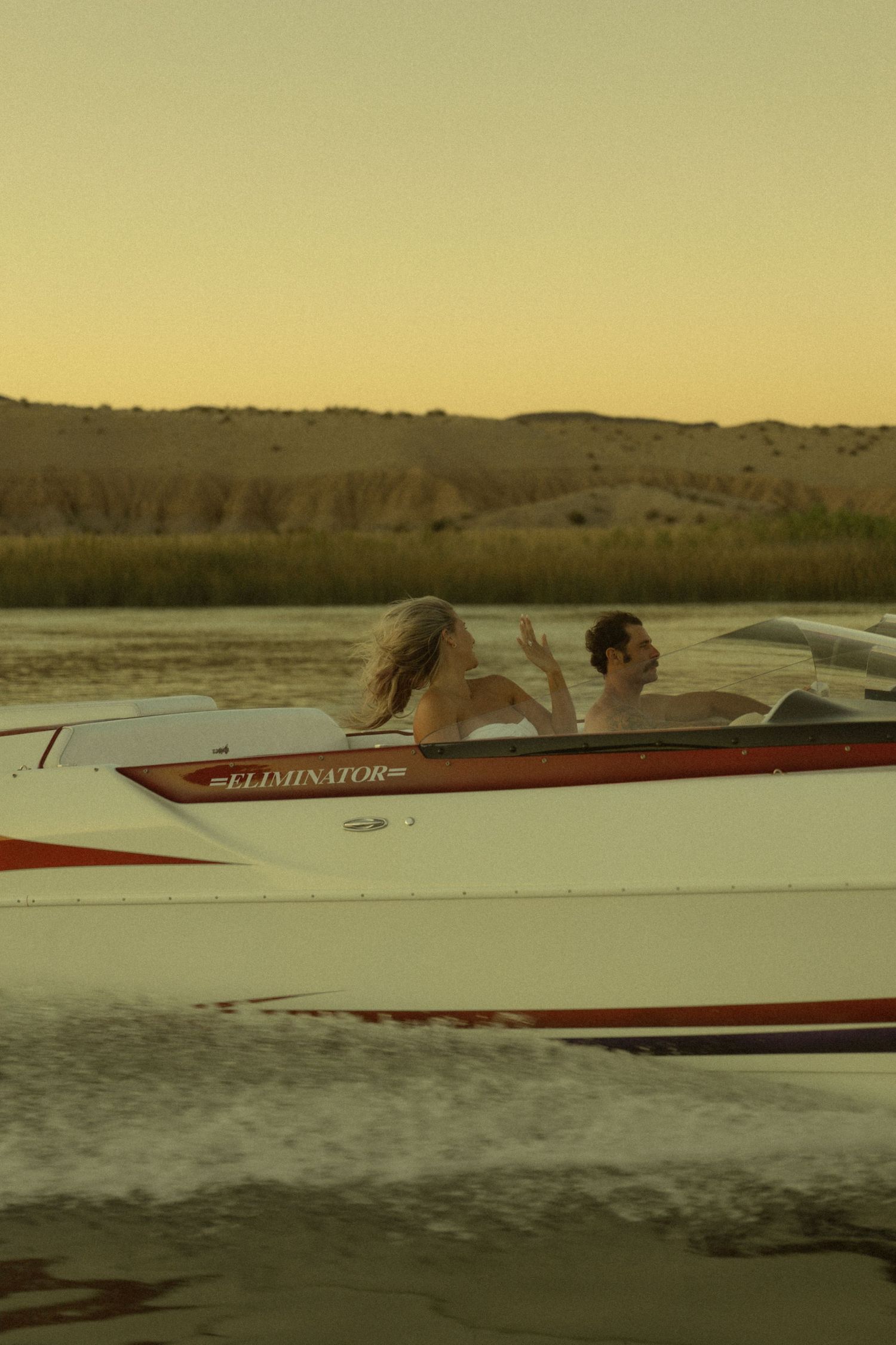 Couple riding in a white and red Eliminator speedboat on a river at sunset.