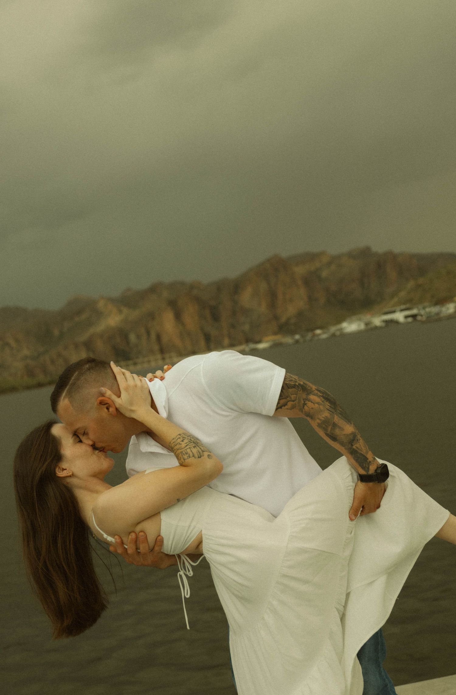 Romantic couple embraces in a passionate kiss pose against a scenic mountain backdrop during sunset.