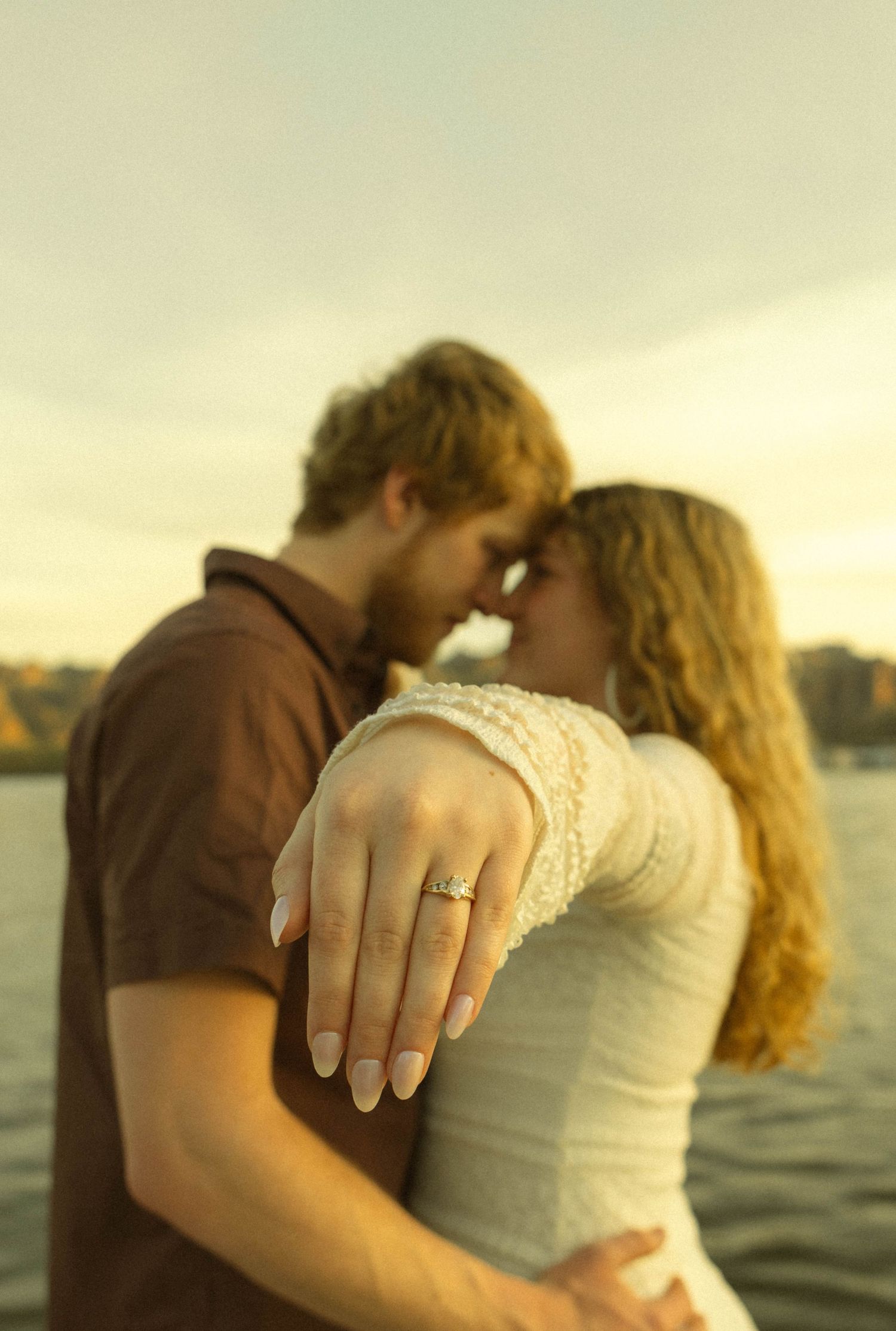 A couple embraces while showing off an engagement ring against a waterfront sunset view.