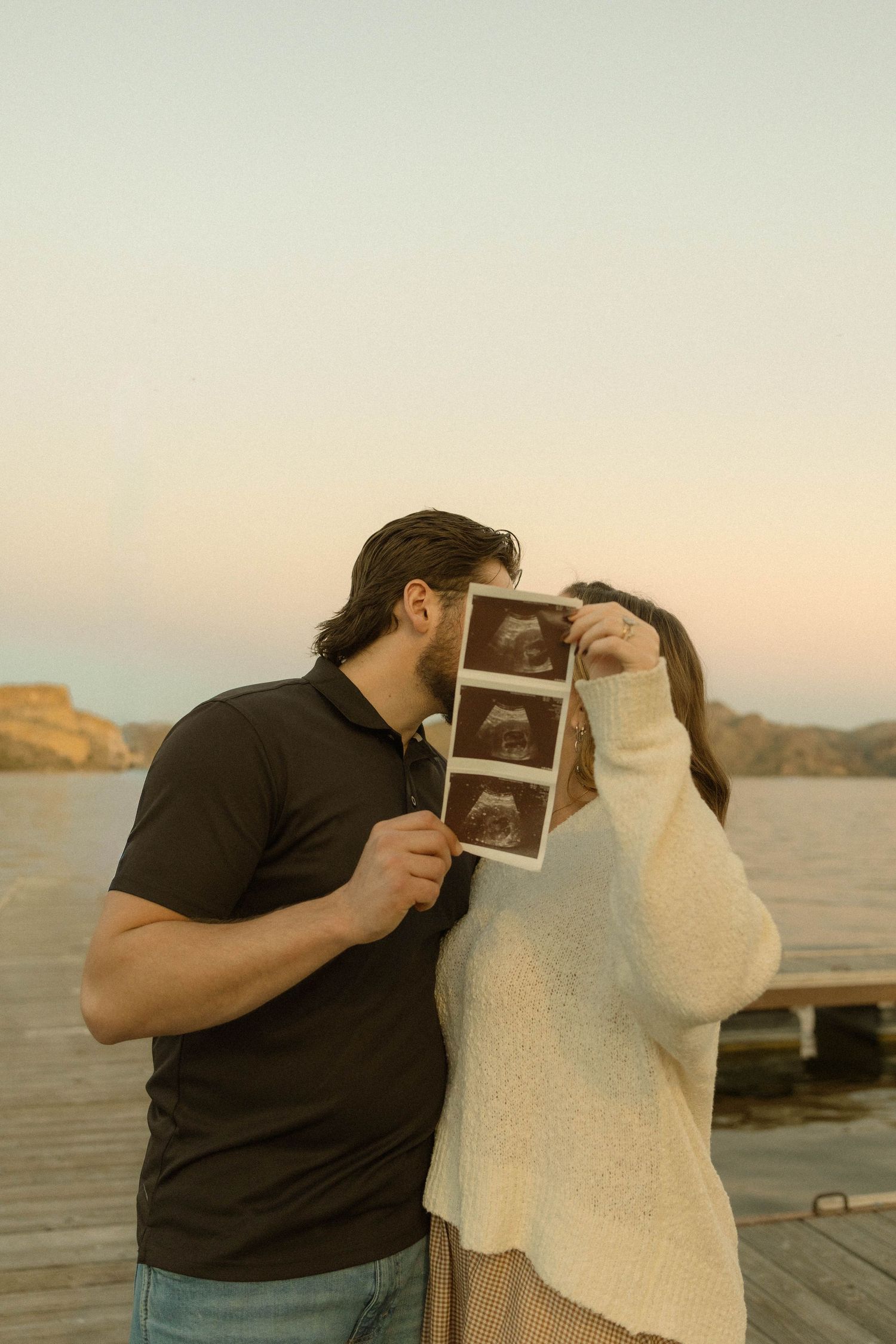 A couple on a dock at sunset sharing an ultrasound photo in a romantic pregnancy announcement series.