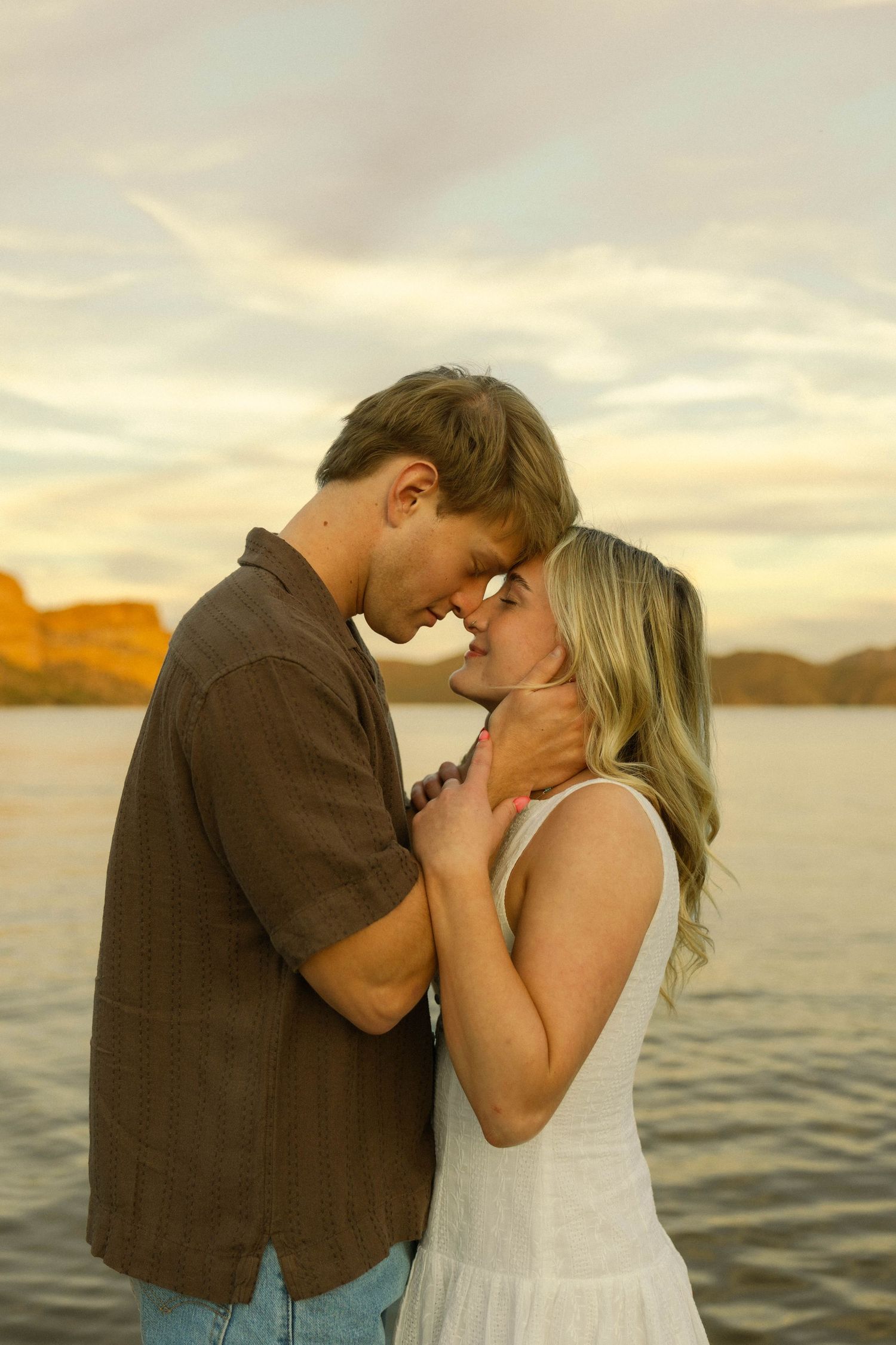 A romantic couple embraces during sunset by a lake with mountains in the background.