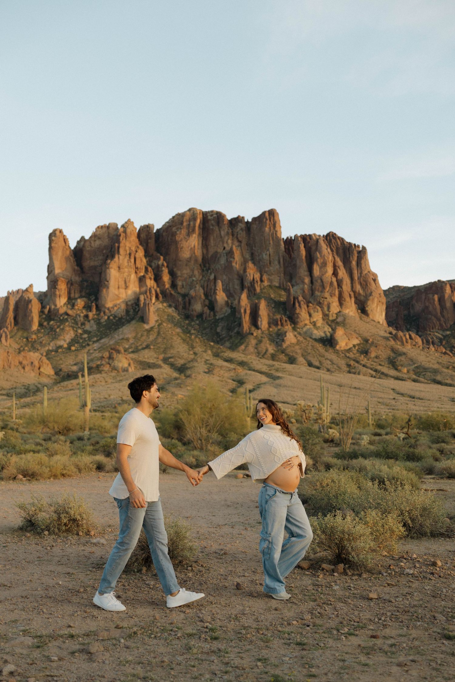 A couple holds hands while walking along a desert trail with towering rock formations in the background.