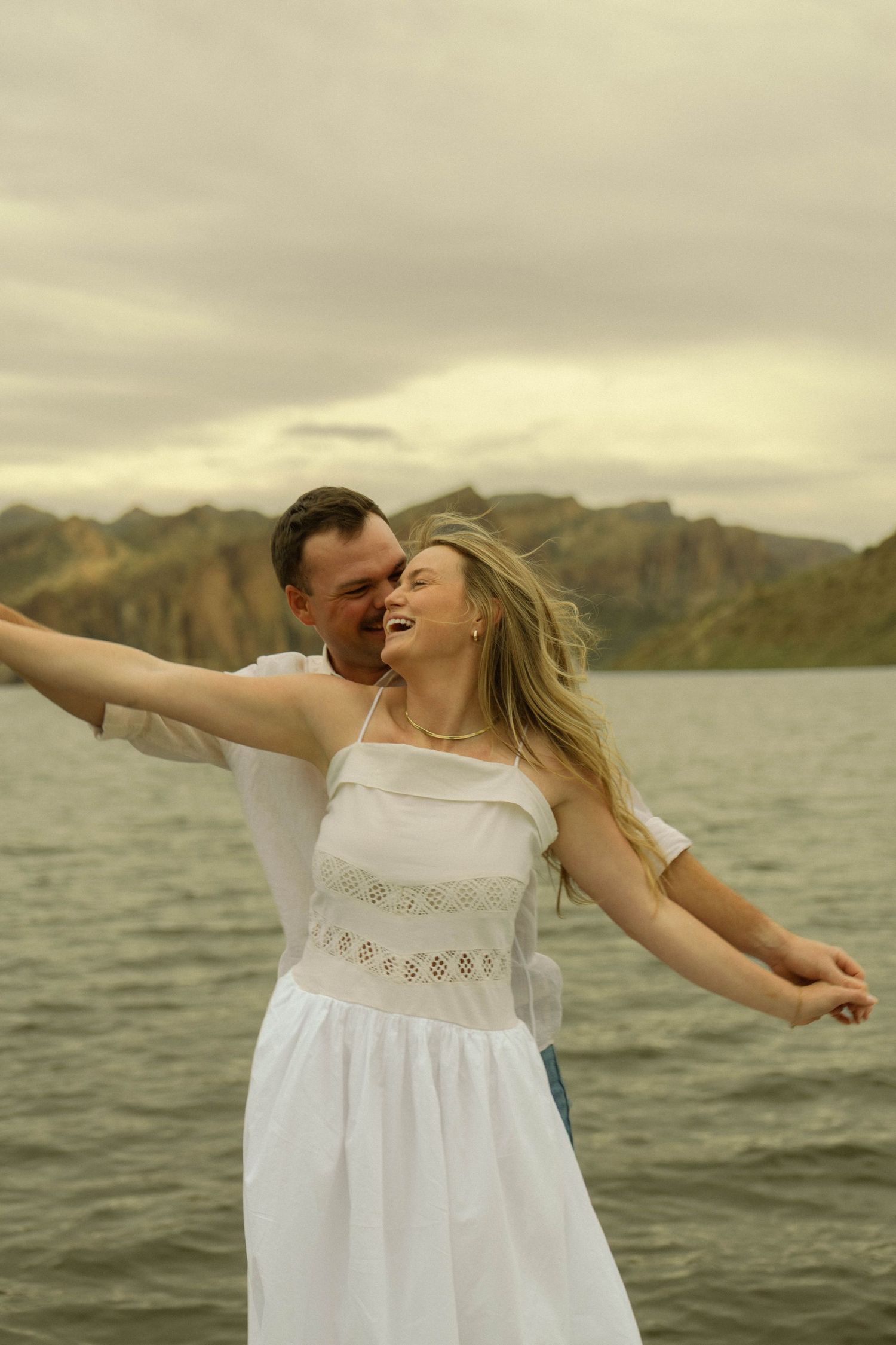 A couple in white clothing dances playfully by a lake with mountainous backdrop.