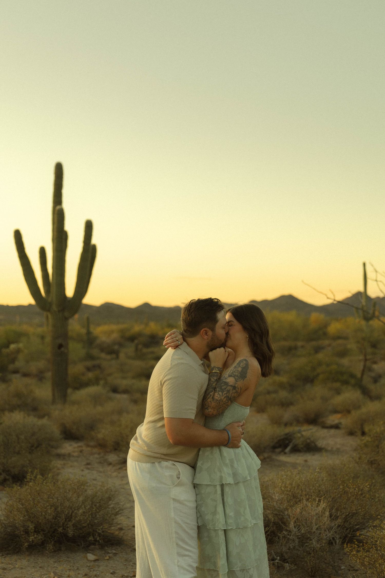 A couple shares a romantic kiss at sunset in the Arizona desert with saguaro cacti in the background.