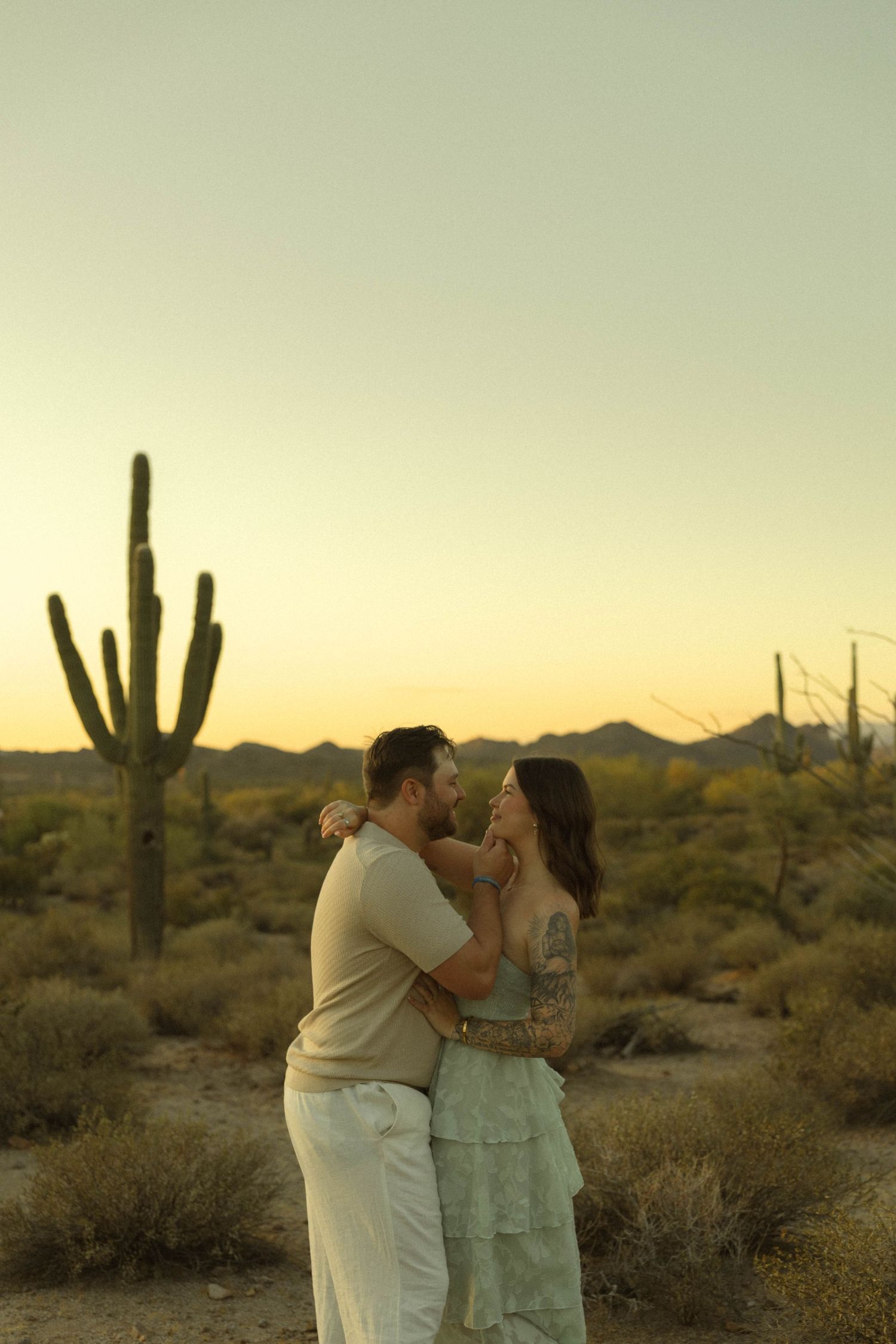 Couple embracing at sunset in Arizona desert with saguaro cacti and mountain backdrop.