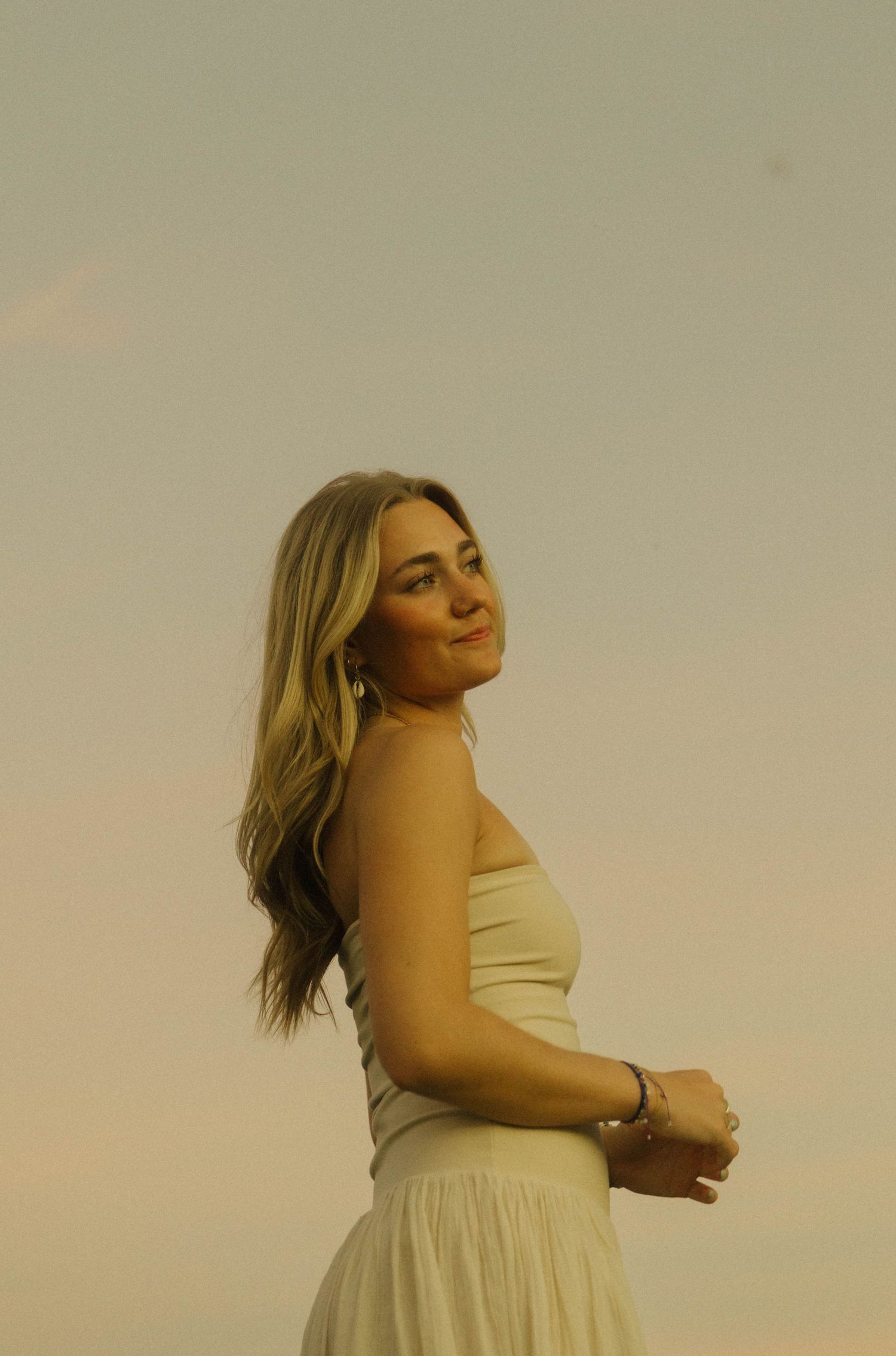 Blonde woman in white strapless dress posing against a soft sunset sky outdoors.