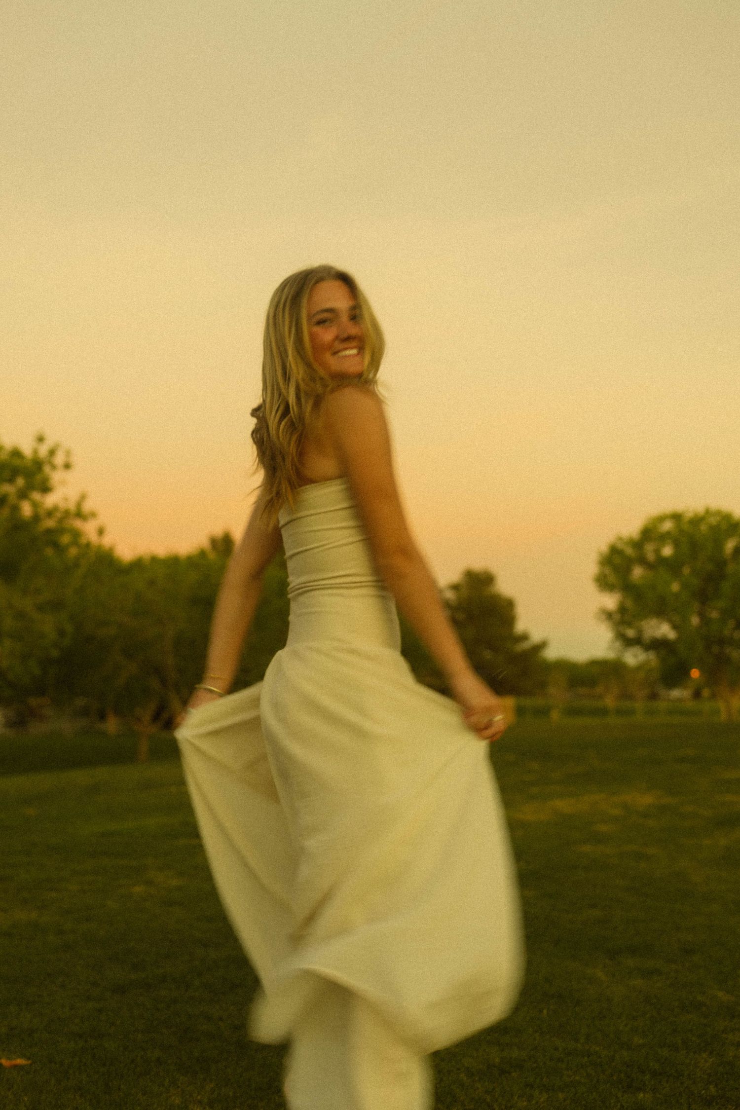 A smiling blonde woman twirls in a white dress at golden hour sunset in a lush green park.