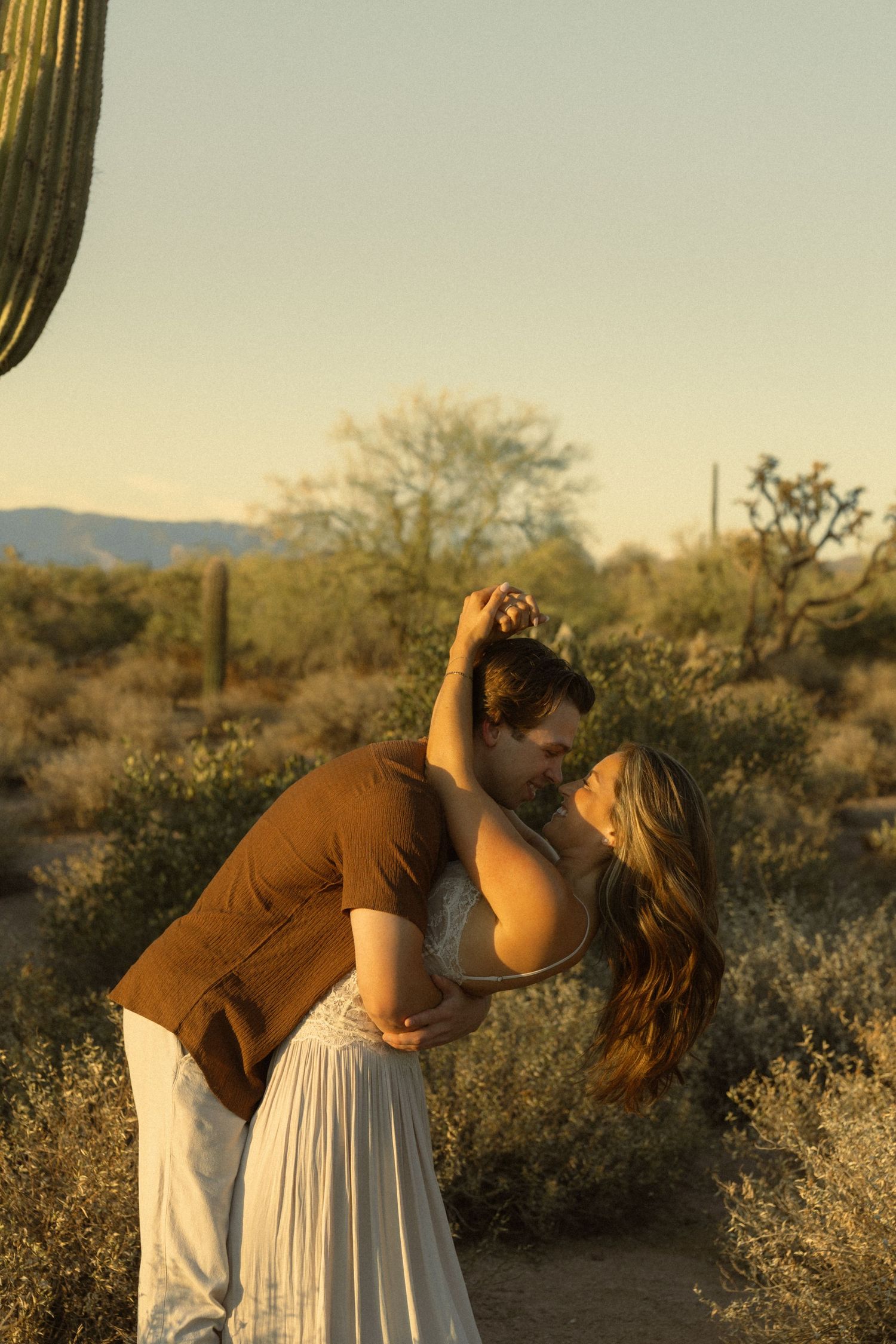 Couple dancing romantically in Arizona desert at golden hour with saguaro cacti and mountains in background.