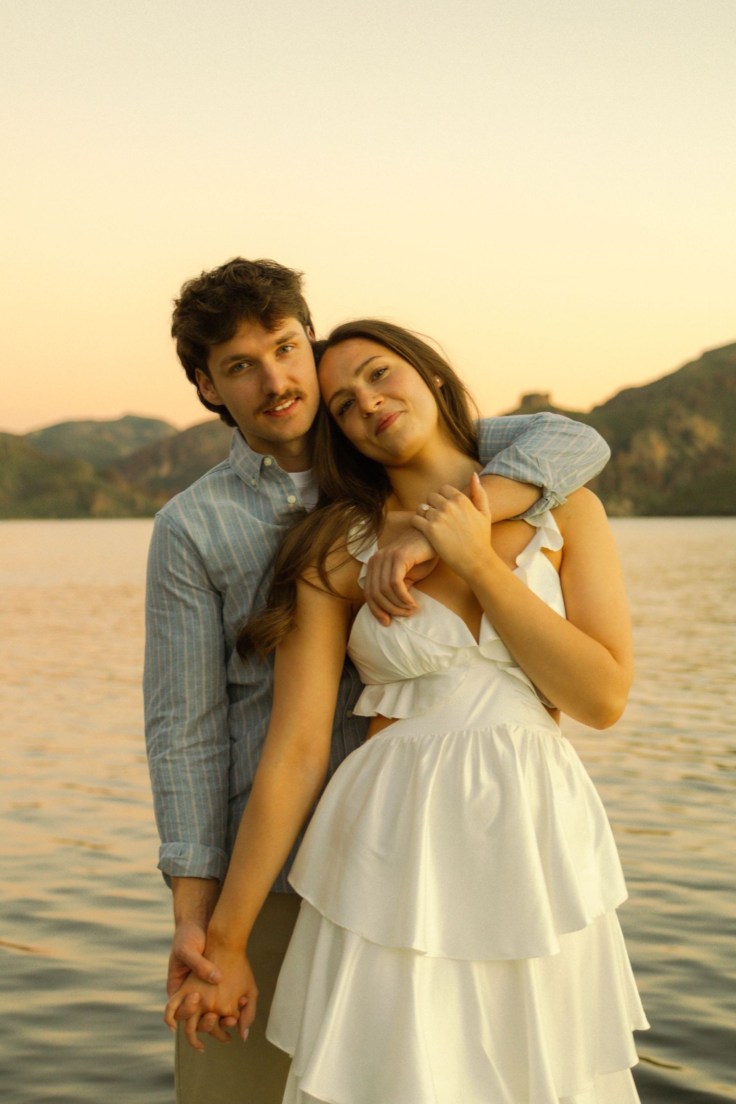 A tender moment between a couple in white and blue clothing against a sunset lake backdrop.