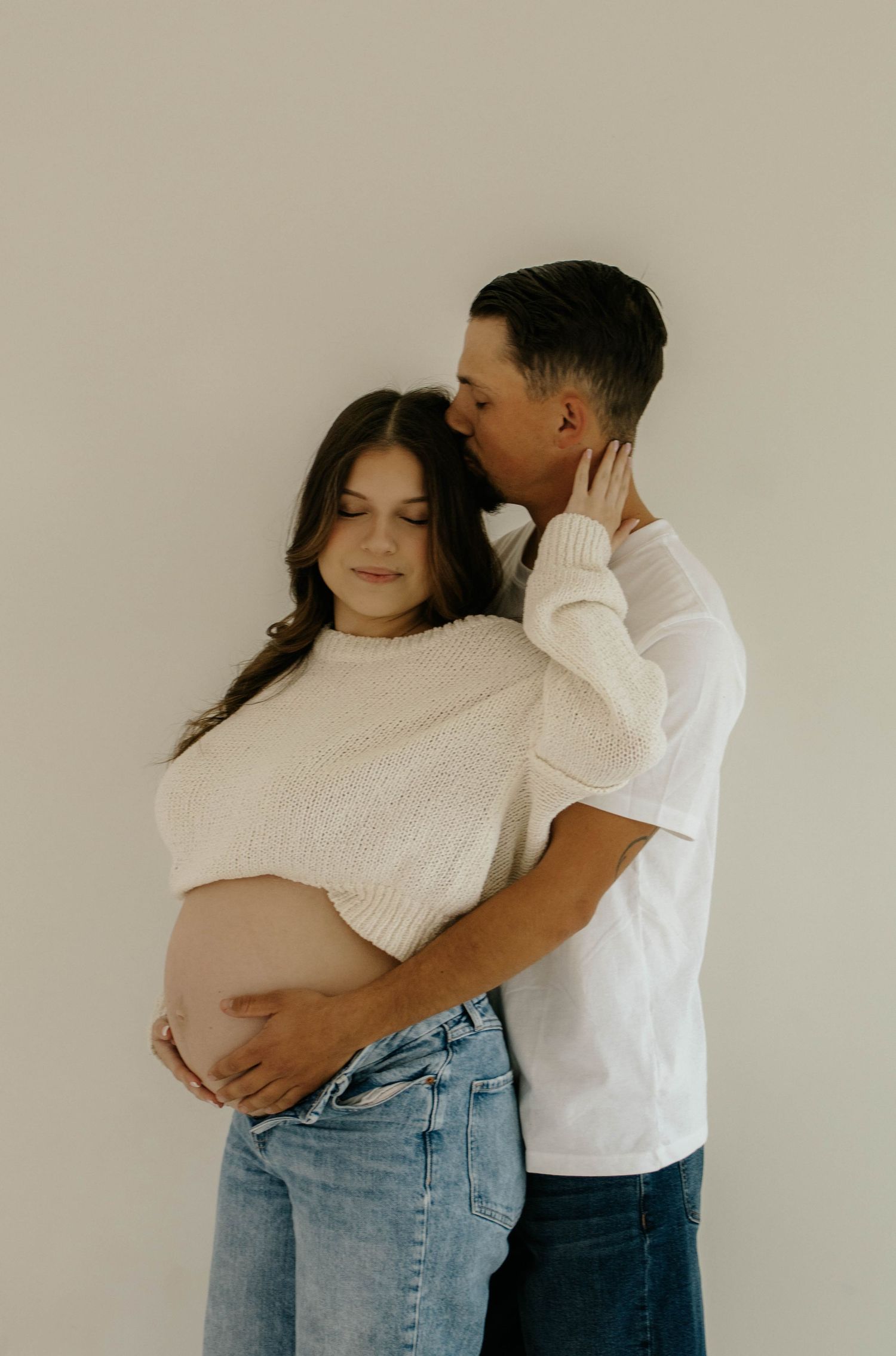 A series of intimate maternity photos showing a couple embracing in white and denim against a neutral background.