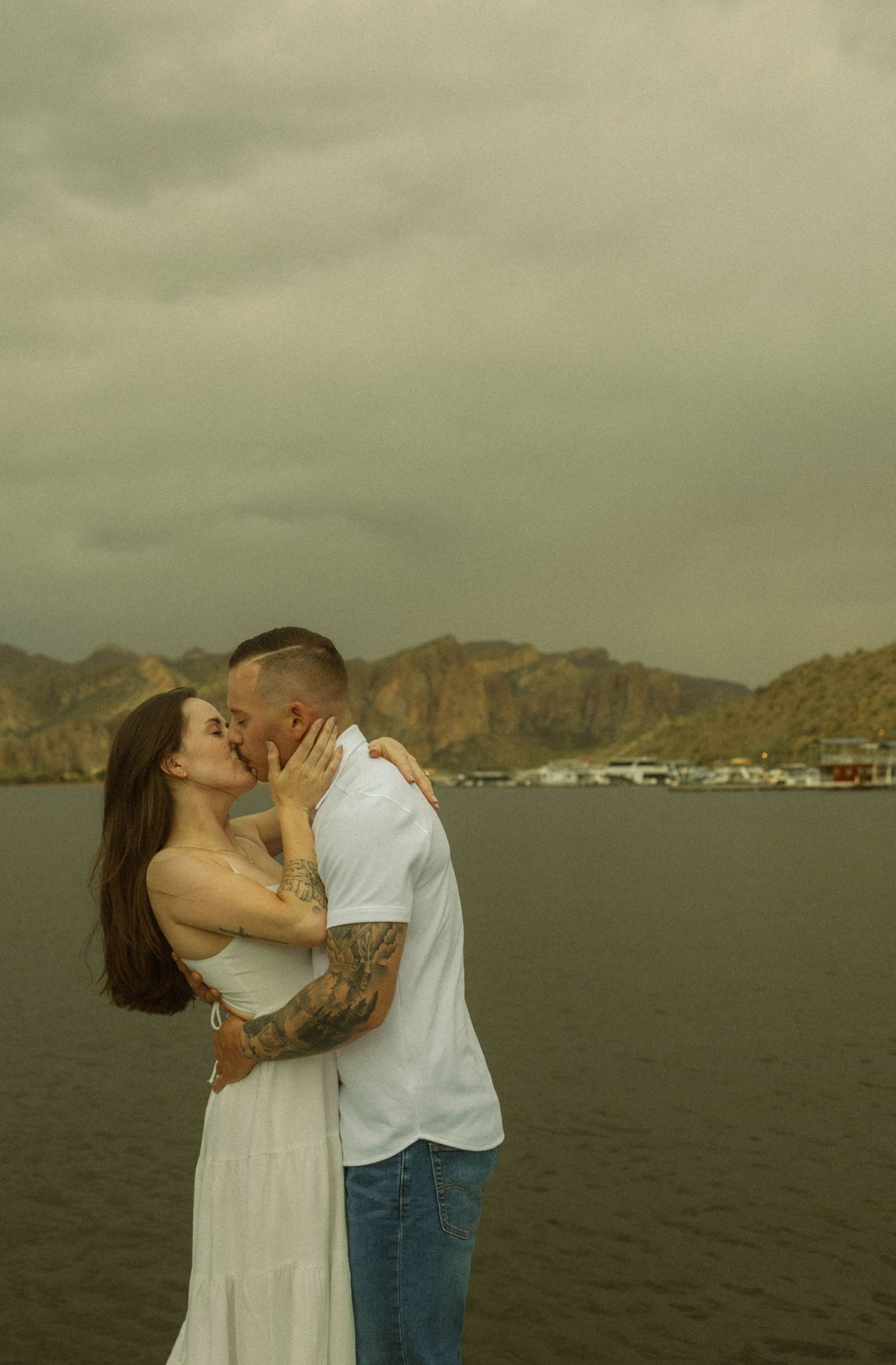 A couple shares a romantic kiss against a scenic mountain backdrop with water and cloudy skies.