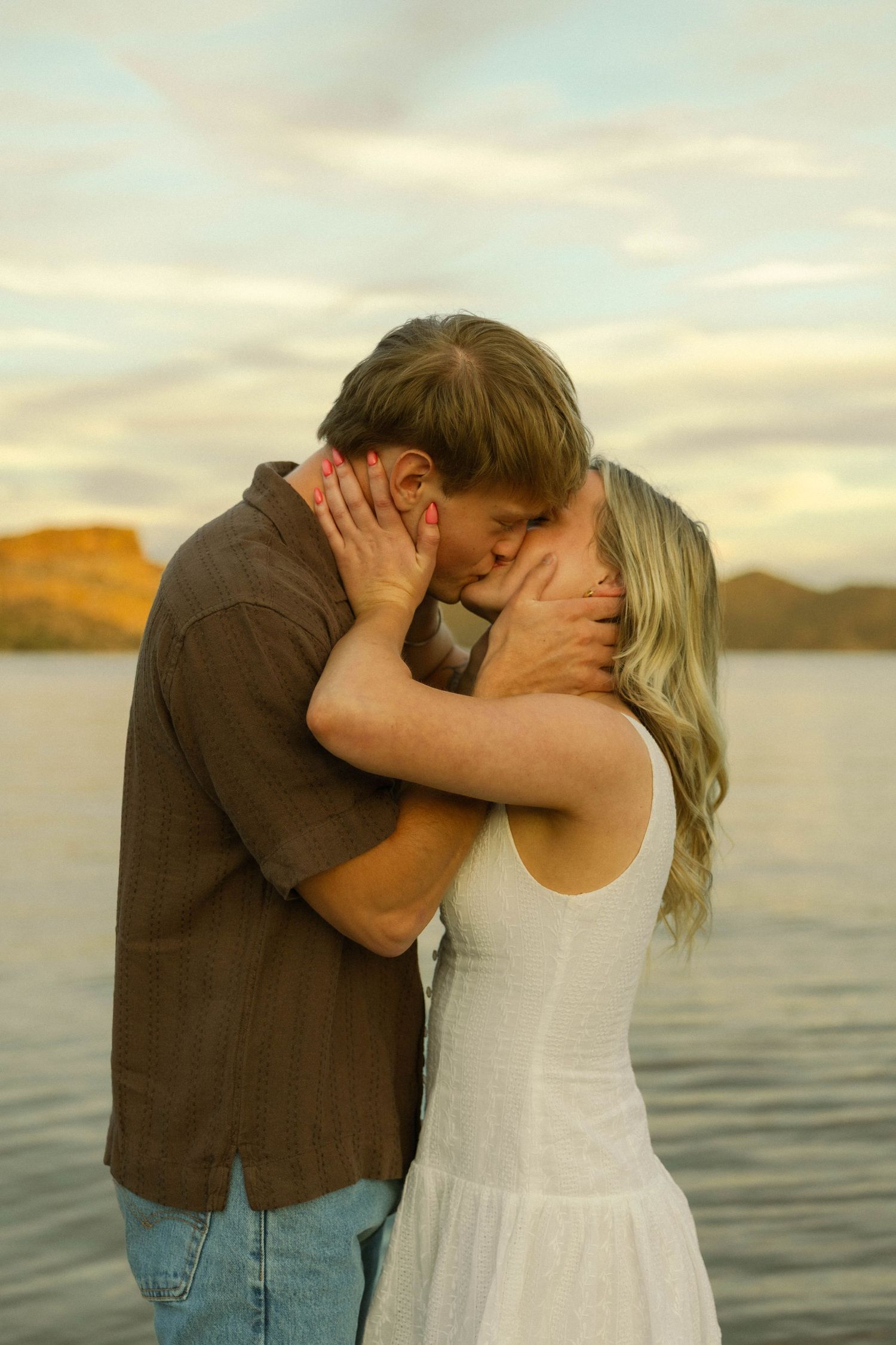 A romantic couple shares a tender kiss at sunset by the water with mountains in the background.