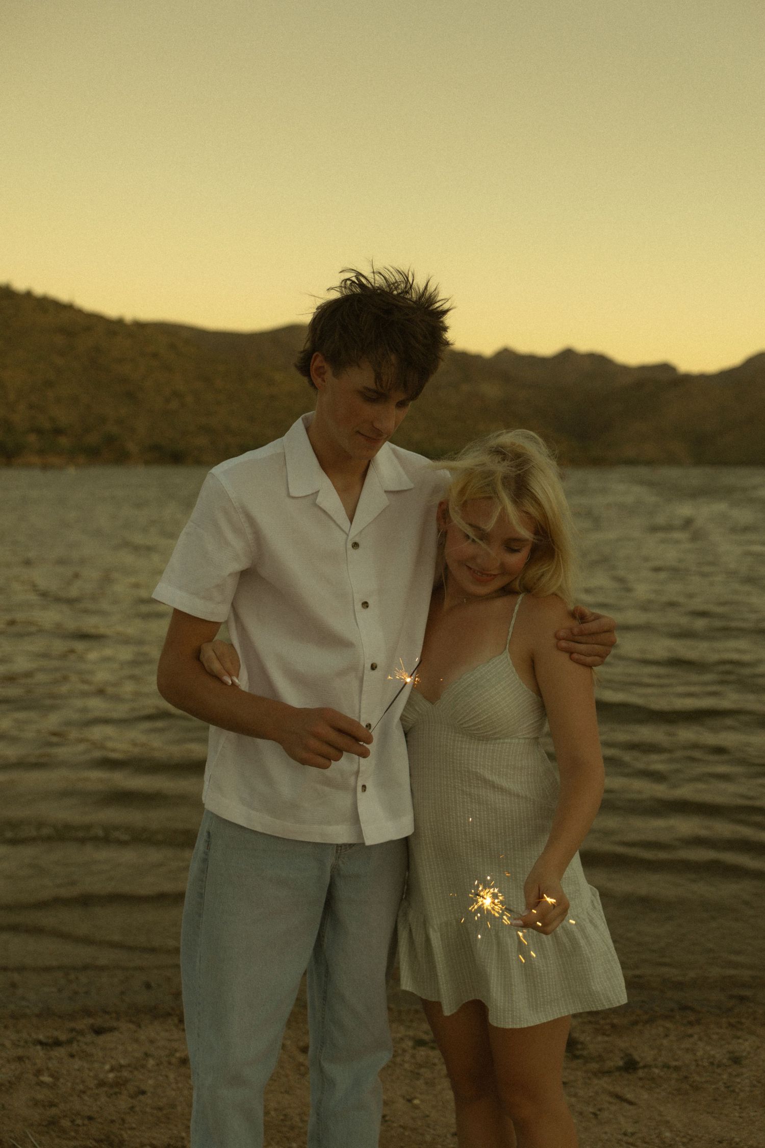A young couple holds sparklers at sunset by a lake, the man in white shirt embracing the blonde woman in a white dress.