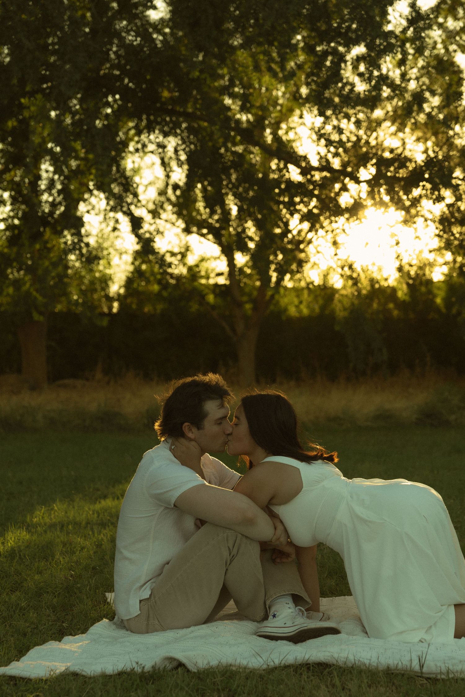 Couple sharing a romantic kiss on a blanket in a lush green park during golden hour sunset.
