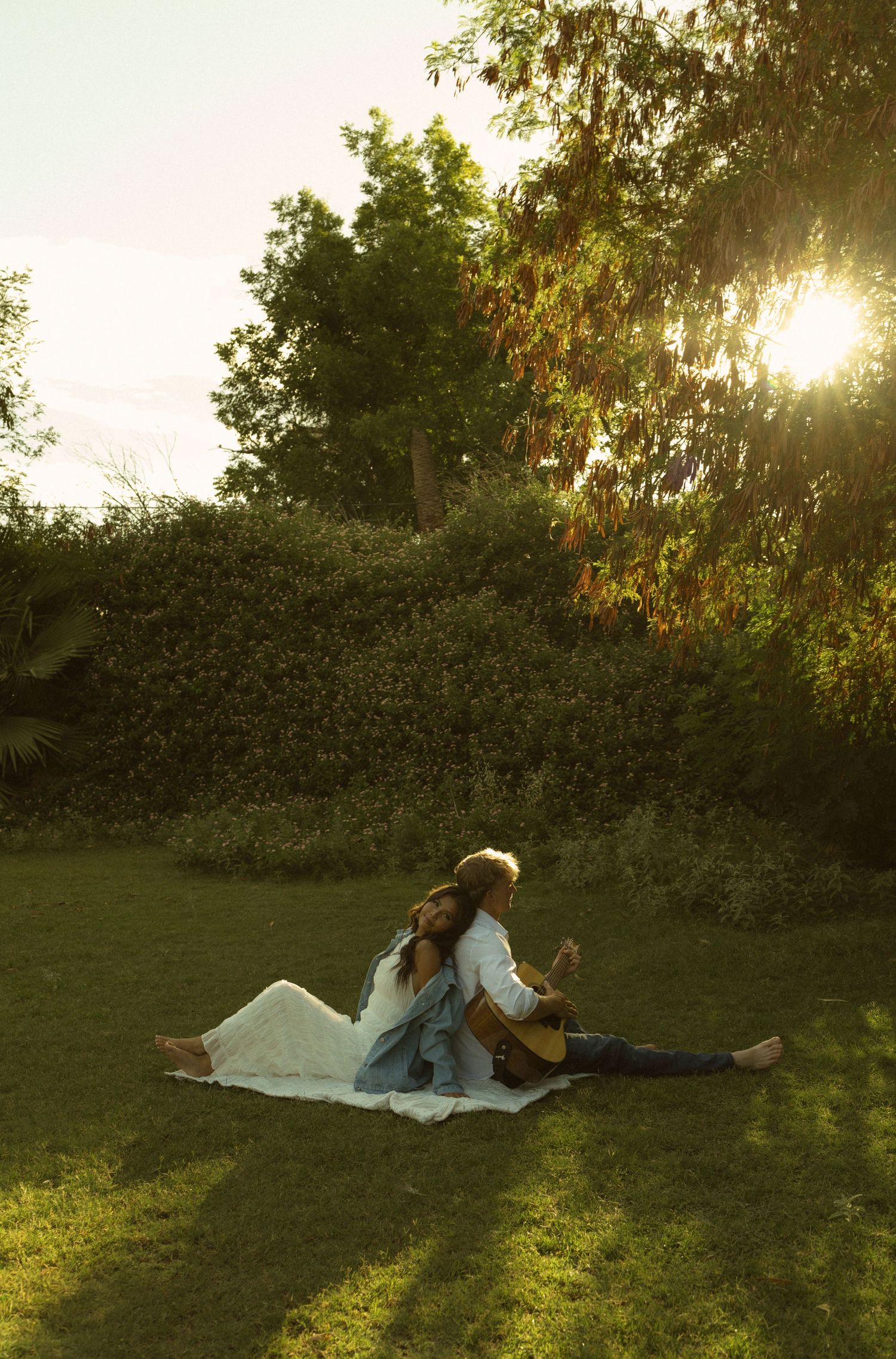 Couple sitting on blanket in sunny garden, one playing guitar, surrounded by lush green trees at golden hour.