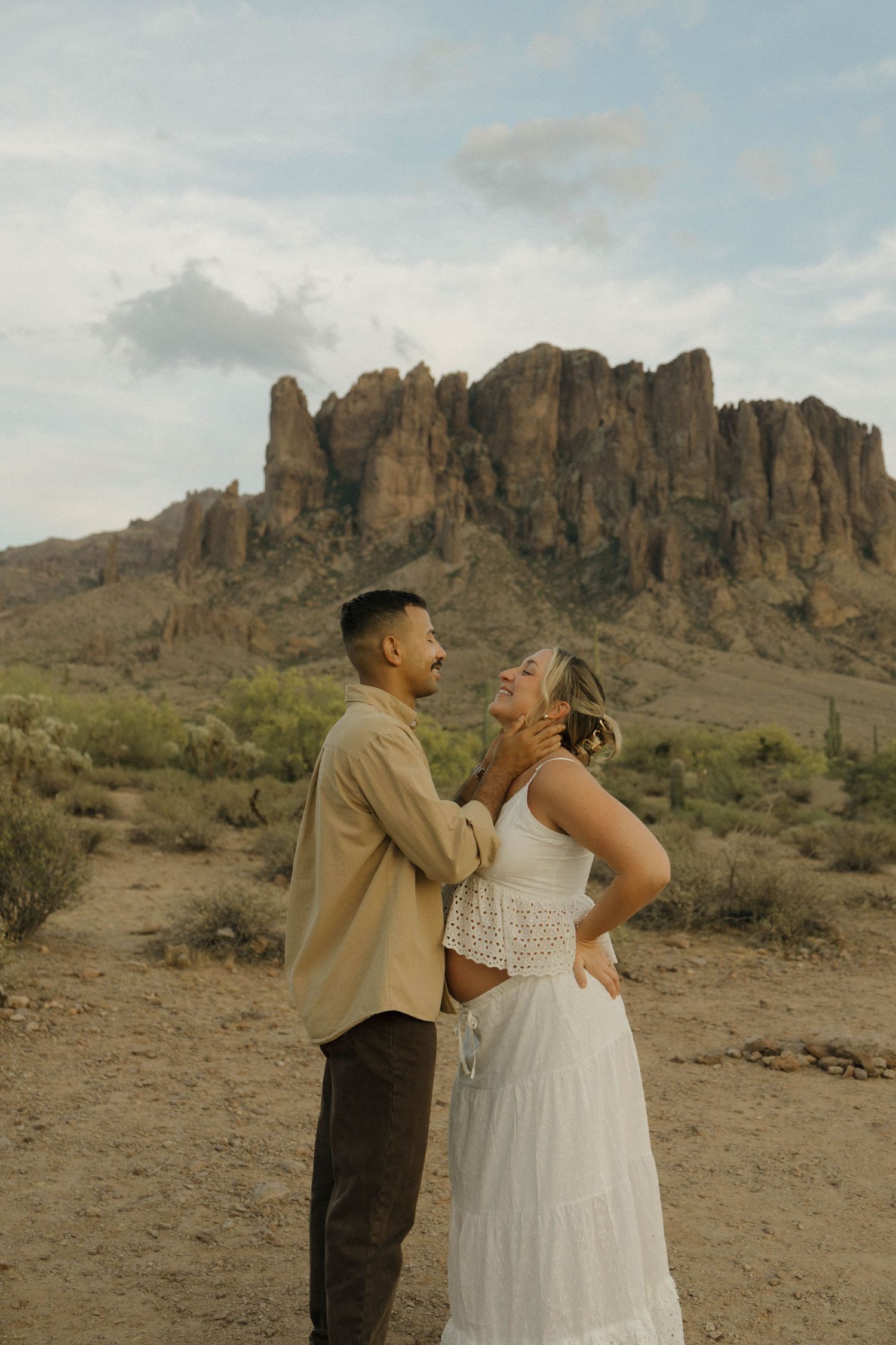 Couple sharing a romantic moment in the Arizona desert with Superstition Mountains backdrop, woman in white dress laughing.