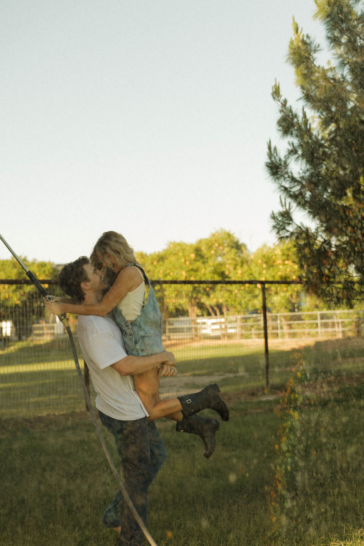 A couple shares a romantic kiss as the man lifts the woman in his arms outdoors on a sunny farm.