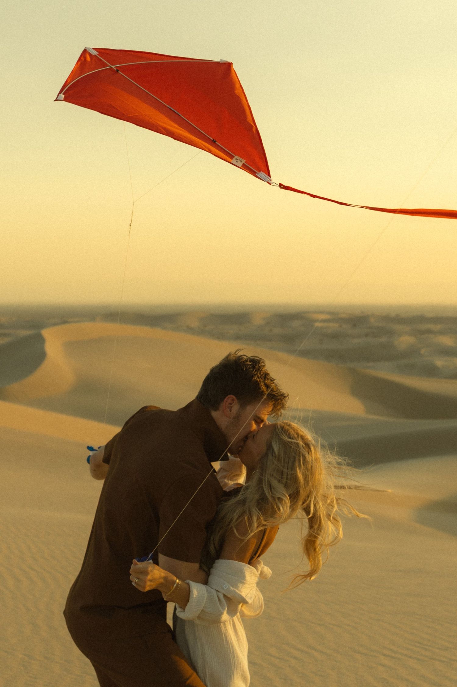 Couple kissing in desert sand dunes at sunset while holding a red kite, romantic outdoor photography.