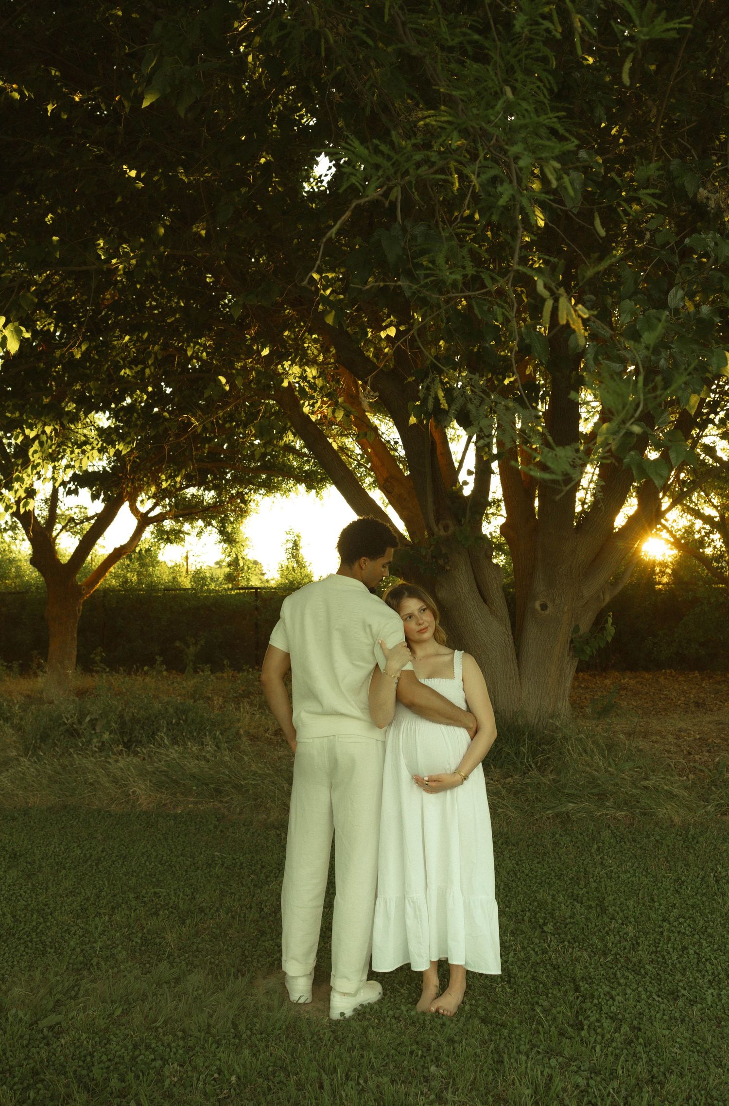 Pregnant woman in white dress with partner under glowing sunset tree during maternity photoshoot.