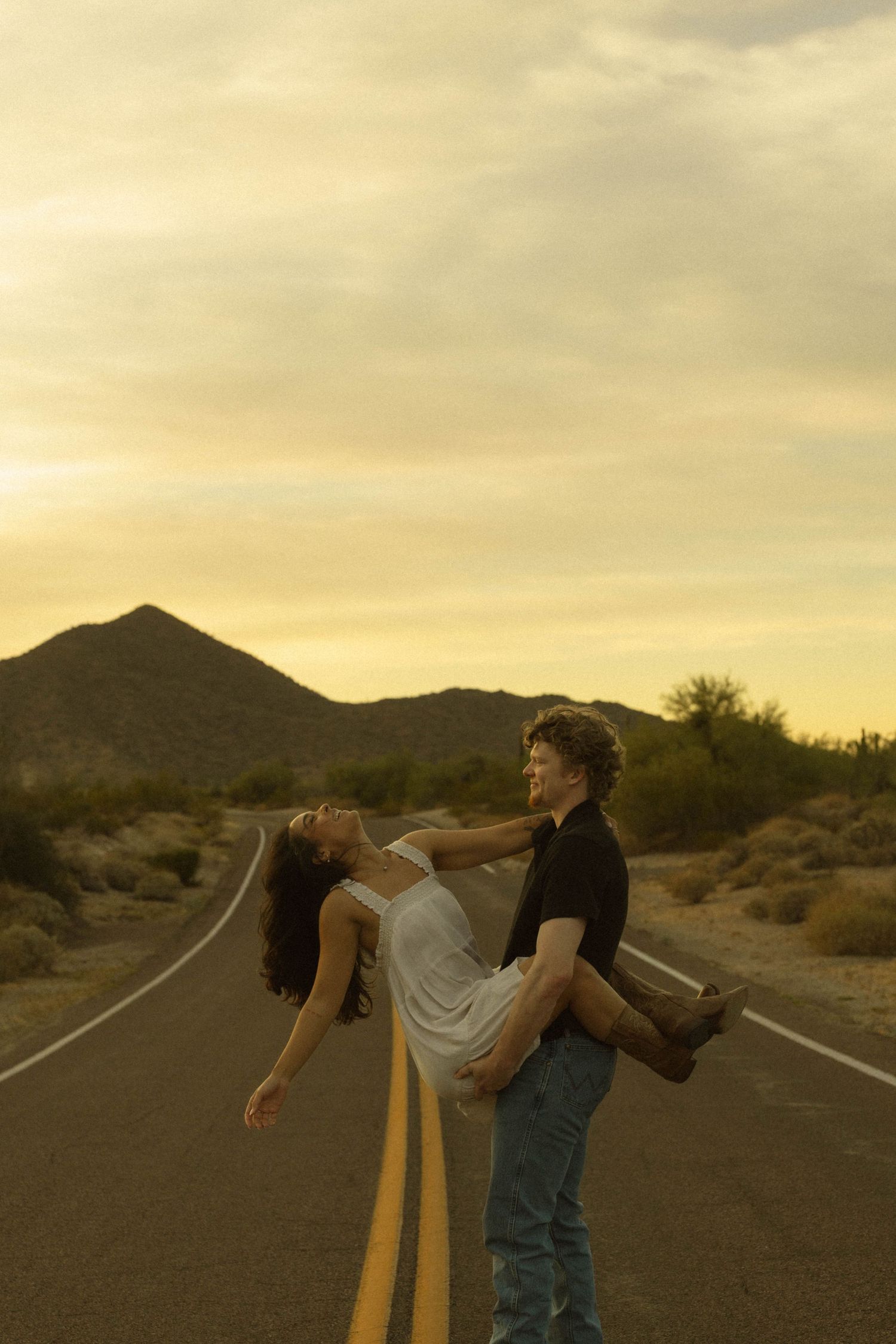 A man lifts a woman in a white dress on a desert road at sunset with mountains in the background.