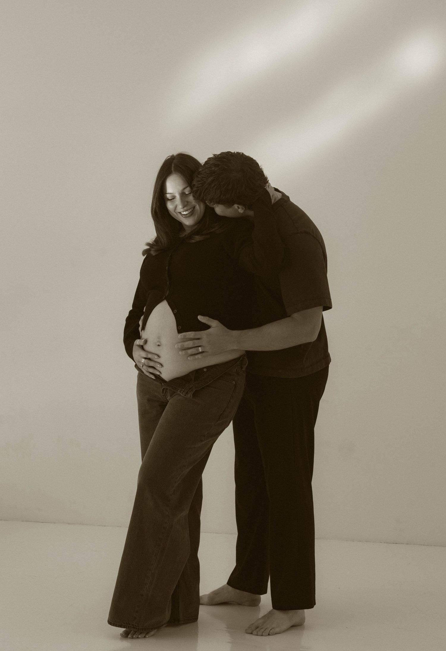 A black and white artistic maternity photo showing a tender embrace against a light background.