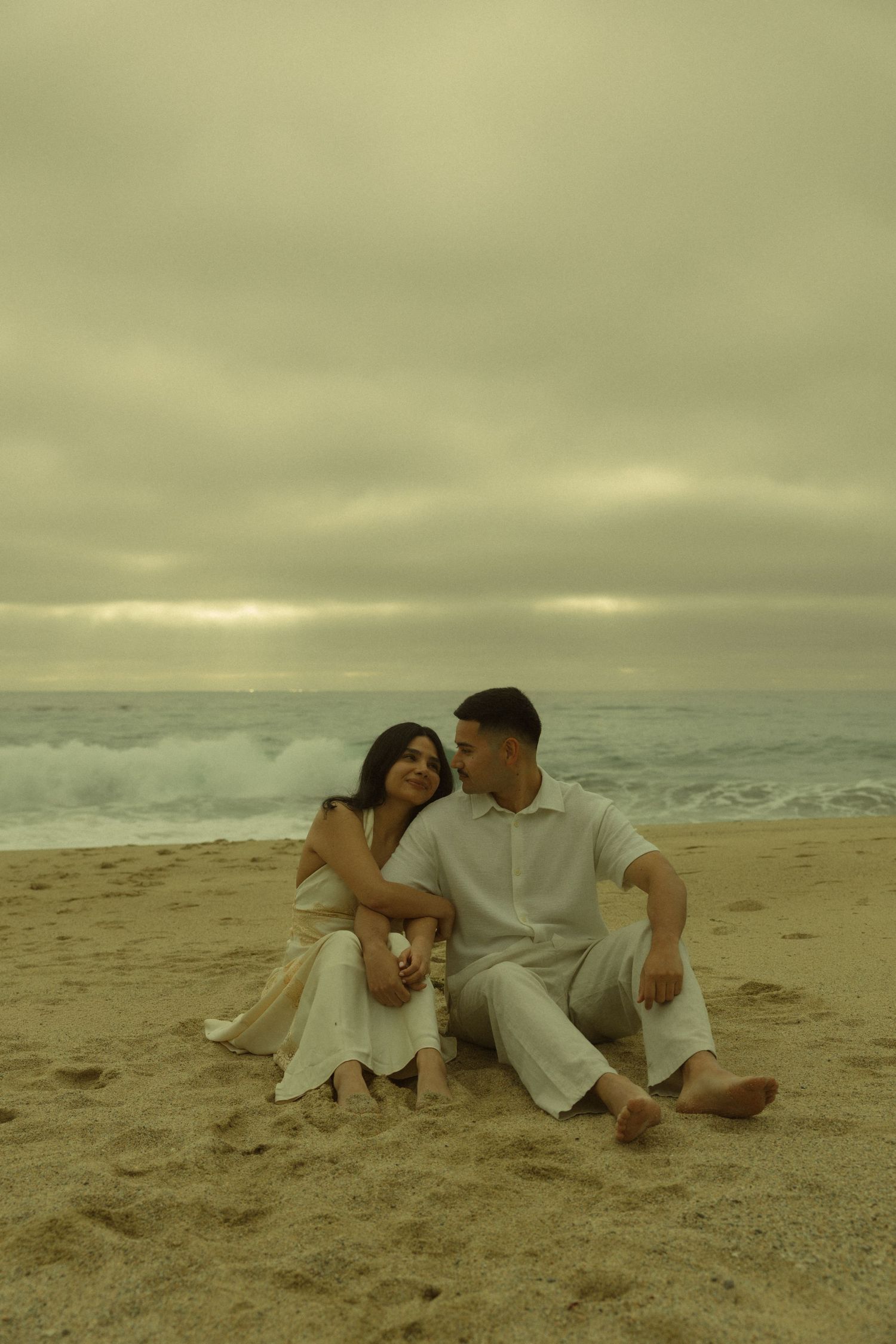 Couple sitting together on sandy beach in white outfits, sharing a romantic moment with ocean waves and cloudy sky.